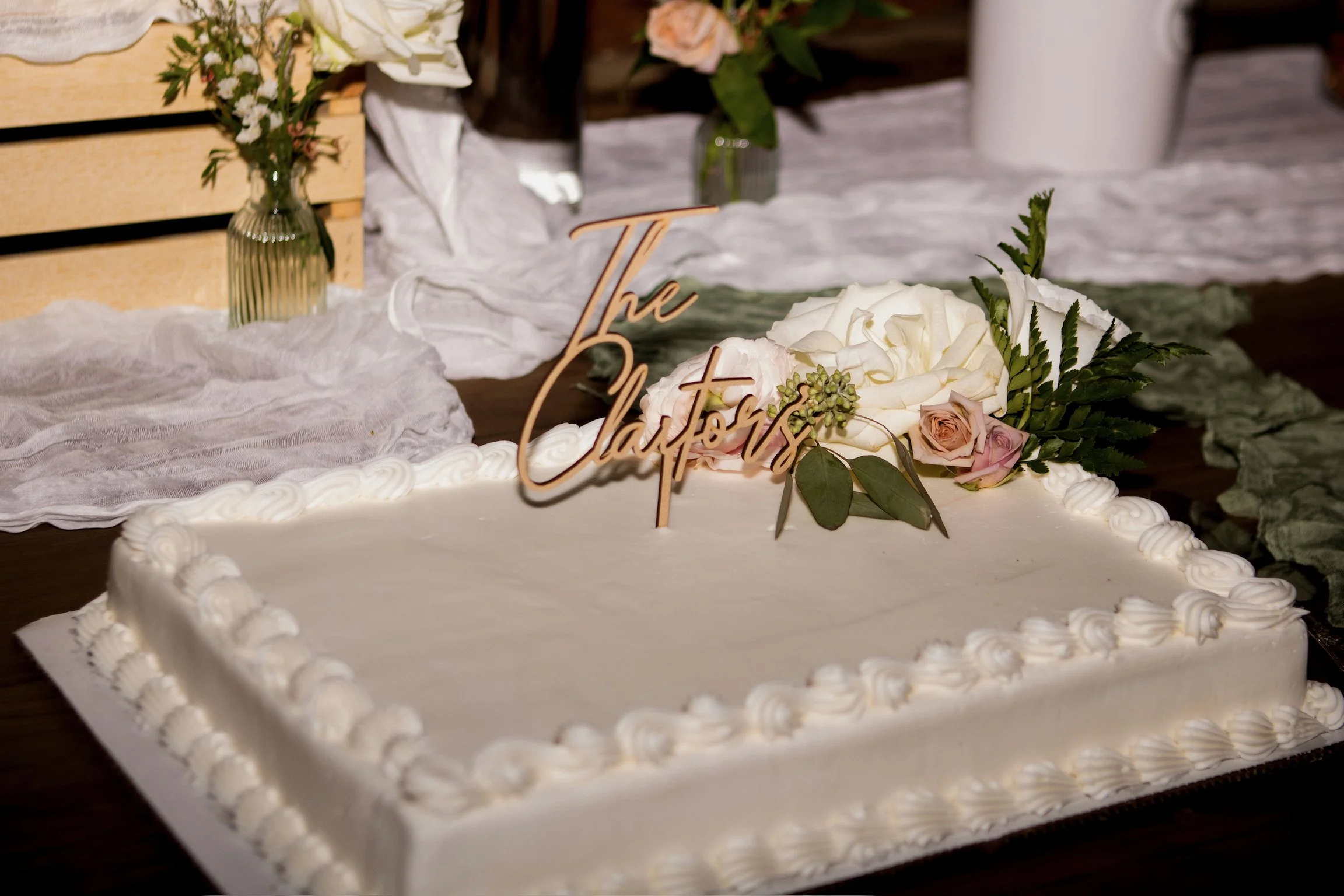 A decorated wedding cake with white icing, topped with pink and white roses, greenery, and a wooden 'true love' sign. In the background, there are vases with flowers and a rustic wooden display.