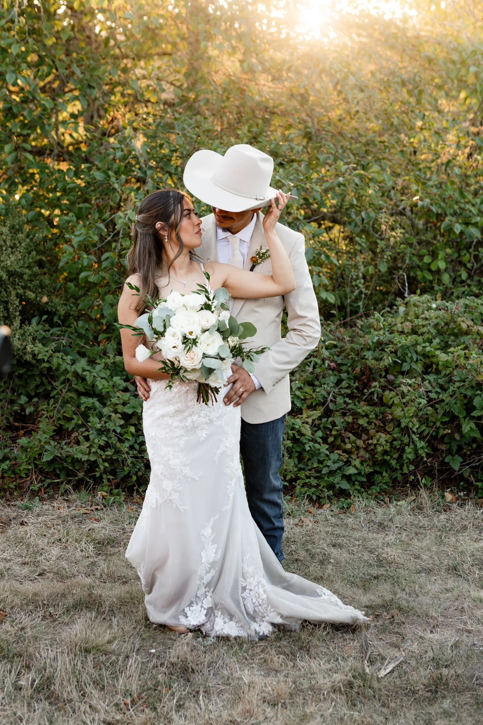 A bride and groom standing outdoors against a backdrop of lush green foliage and sunlight, with the bride holding a bouquet of white flowers and greenery, and the groom wearing a beige suit and white hat, sharing an intimate moment.