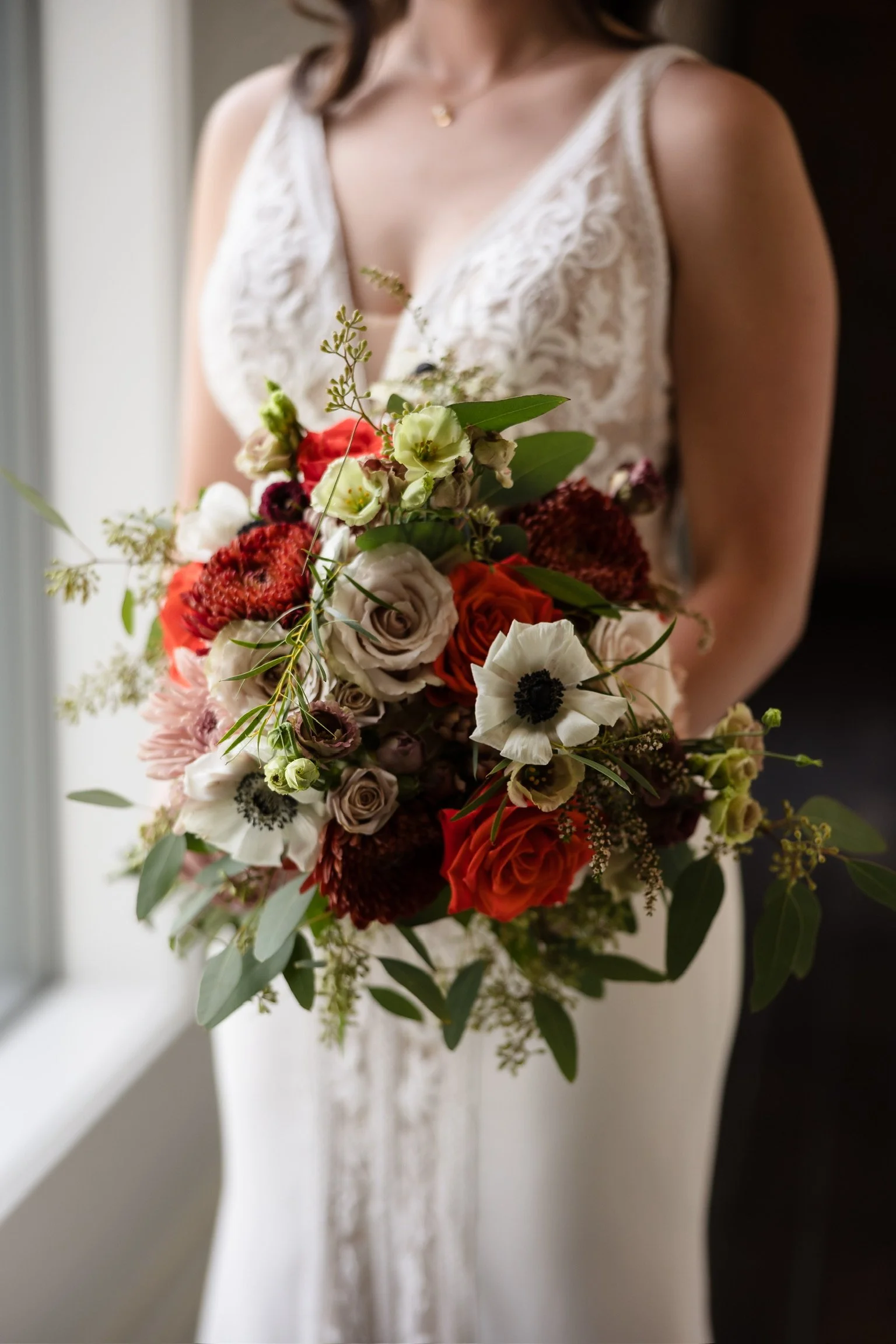 A bride in a lace wedding dress holding a large bouquet of colorful flowers, standing near a window.