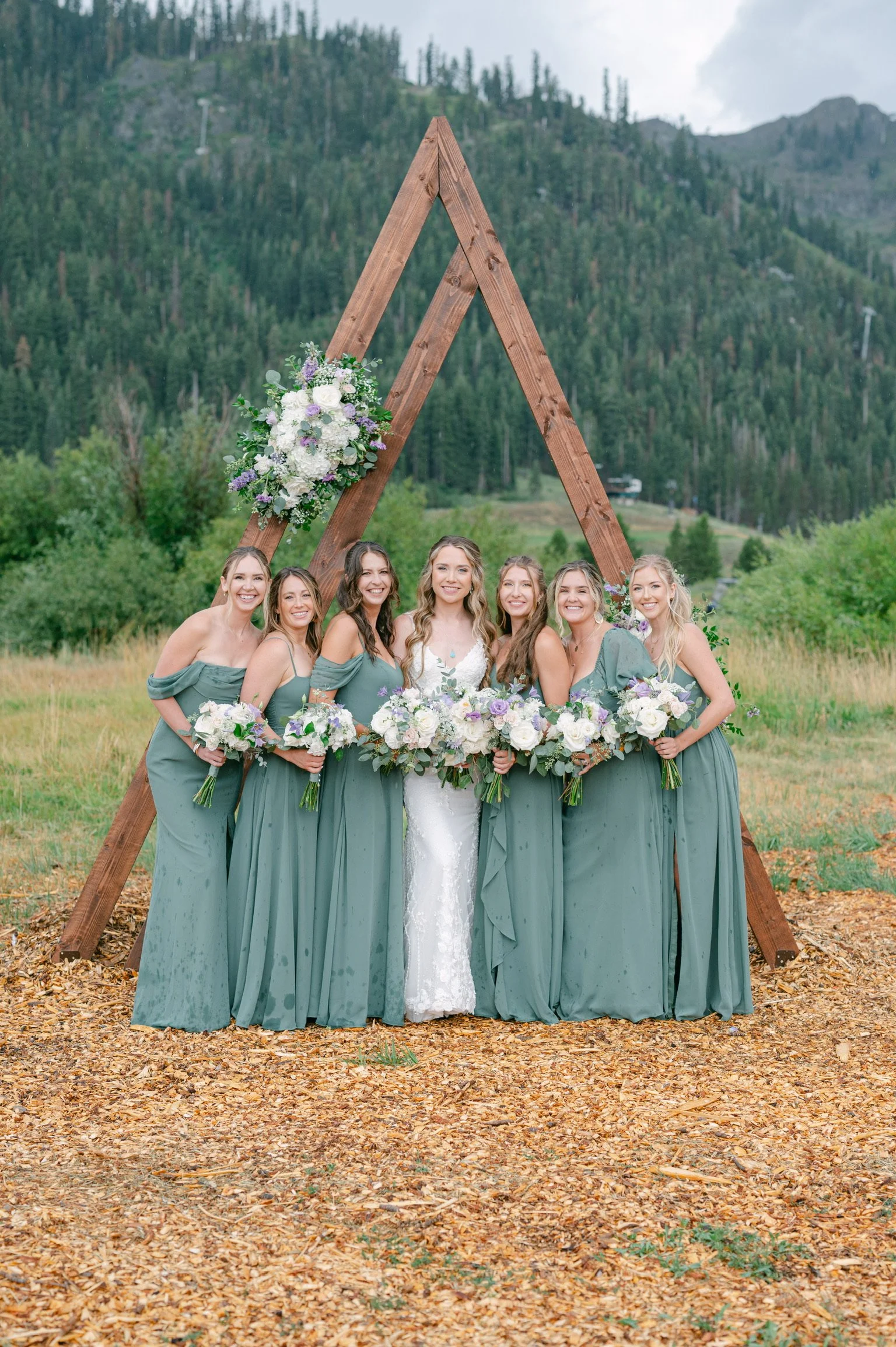 A group of seven women dressed in matching teal bridesmaid dresses and one bride in a white wedding gown, standing outdoors in front of a wooden wedding arch decorated with flowers, surrounded by trees and mountains.