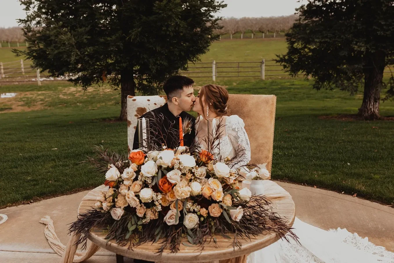 Couple at their wedding, sitting at a table outdoors, kissing, with a large floral arrangement of white and orange roses and greenery in front of them, trees and a farm fence in the background.