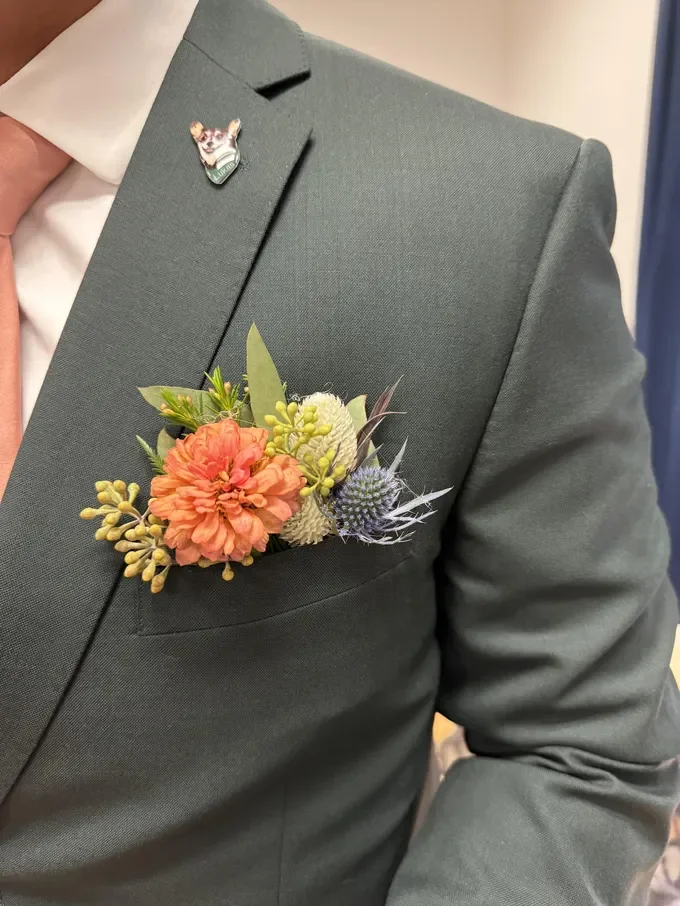 Man wearing a gray suit with a pink shirt and a floral boutonniere on his lapel. The boutonniere features a large orange flower, green leaves, and spiky thistle-like elements.