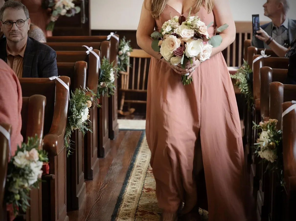 A woman in a pink dress is holding a bouquet of white and blush roses, walking down the aisle of a decorated church during a wedding ceremony. Guests are seated on wooden pews decorated with greenery and white ribbons, and some are taking photos.