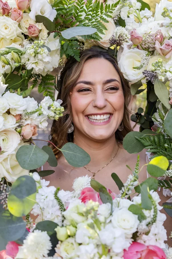 A woman smiling happily among a large arrangement of white and pink flowers and green foliage.