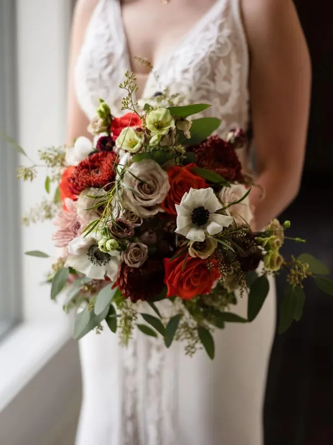 A bride in a lace wedding gown holding a colorful bouquet of roses, anemones, and greenery.