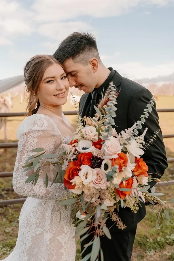 Couple on their wedding day, with the bride holding a large bouquet of flowers, standing outdoors by a fence, smiling and leaning their foreheads together.
