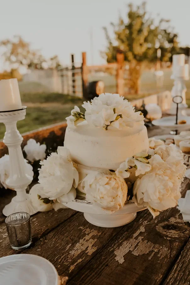 White wedding cake decorated with white flowers on a rustic wooden table with candles and floral arrangements outdoors