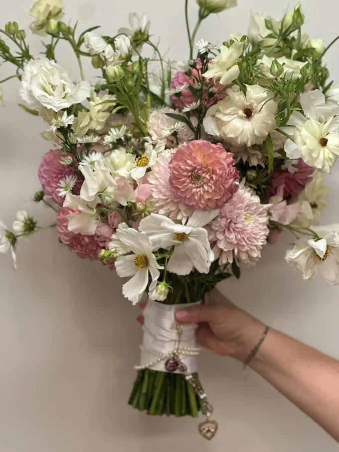 A hand holding a bouquet of pink and white flowers wrapped with white ribbon and decorated with a small heart charm.