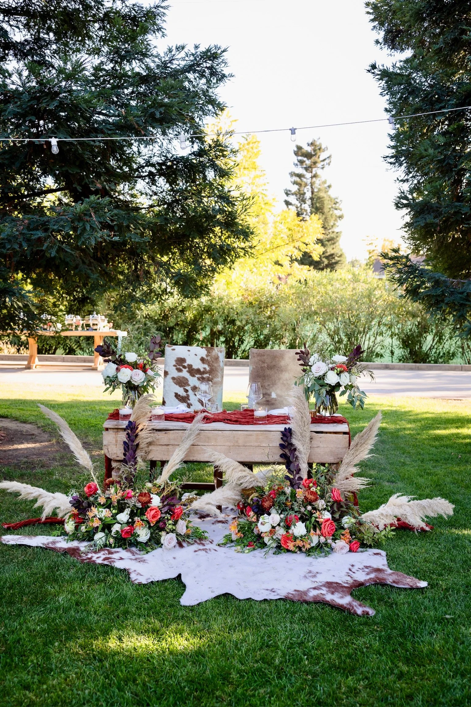 Outdoor table set with floral arrangements, surrounded by pampas grass and a cowhide rug, with trees and string lights in the background.