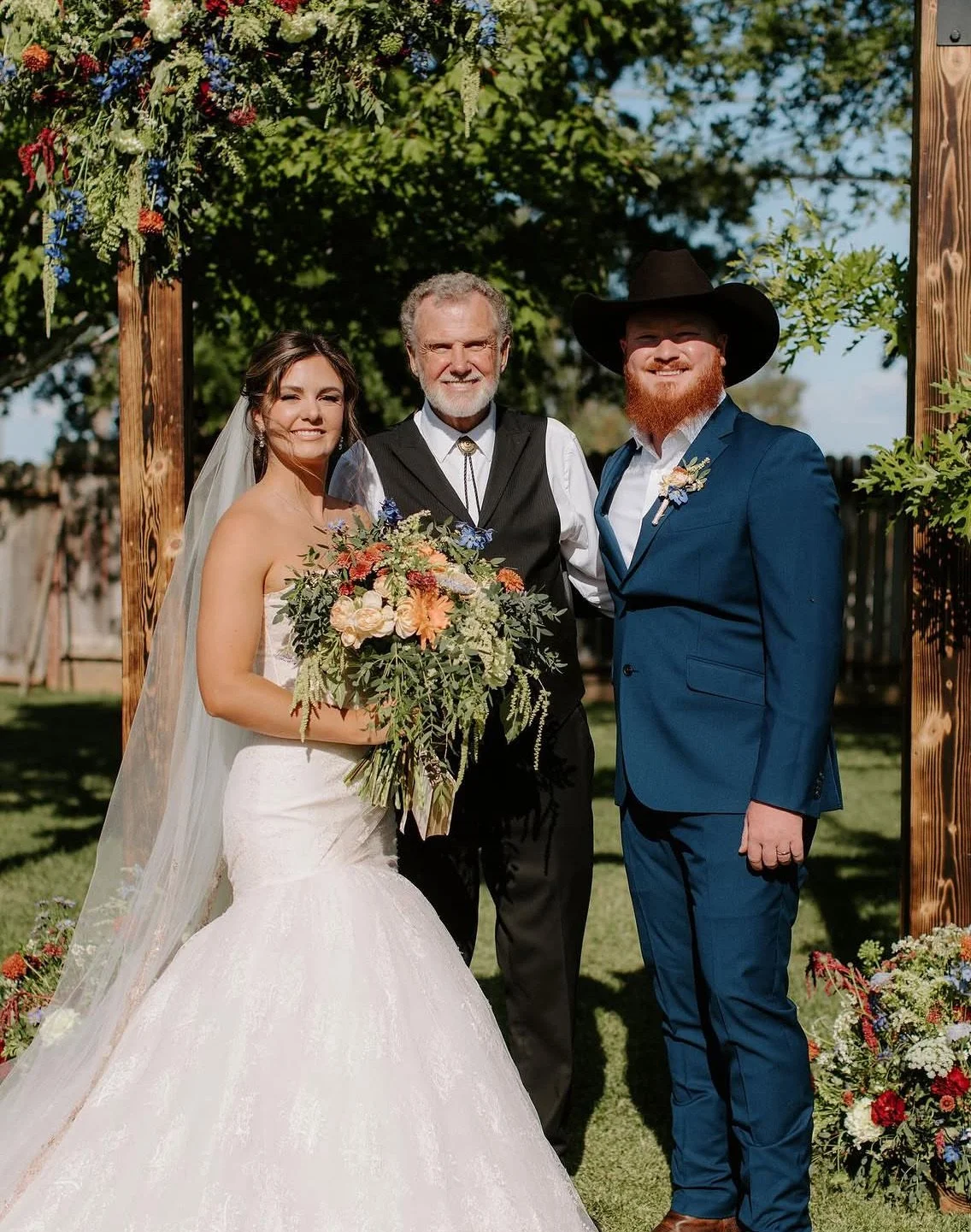 A bride and groom standing with an officiant at a wedding ceremony outdoors. The bride is in a white wedding dress holding a large bouquet, and the groom is in a blue suit with a black wide-brimmed hat. The officiant is in a black vest. They are smil