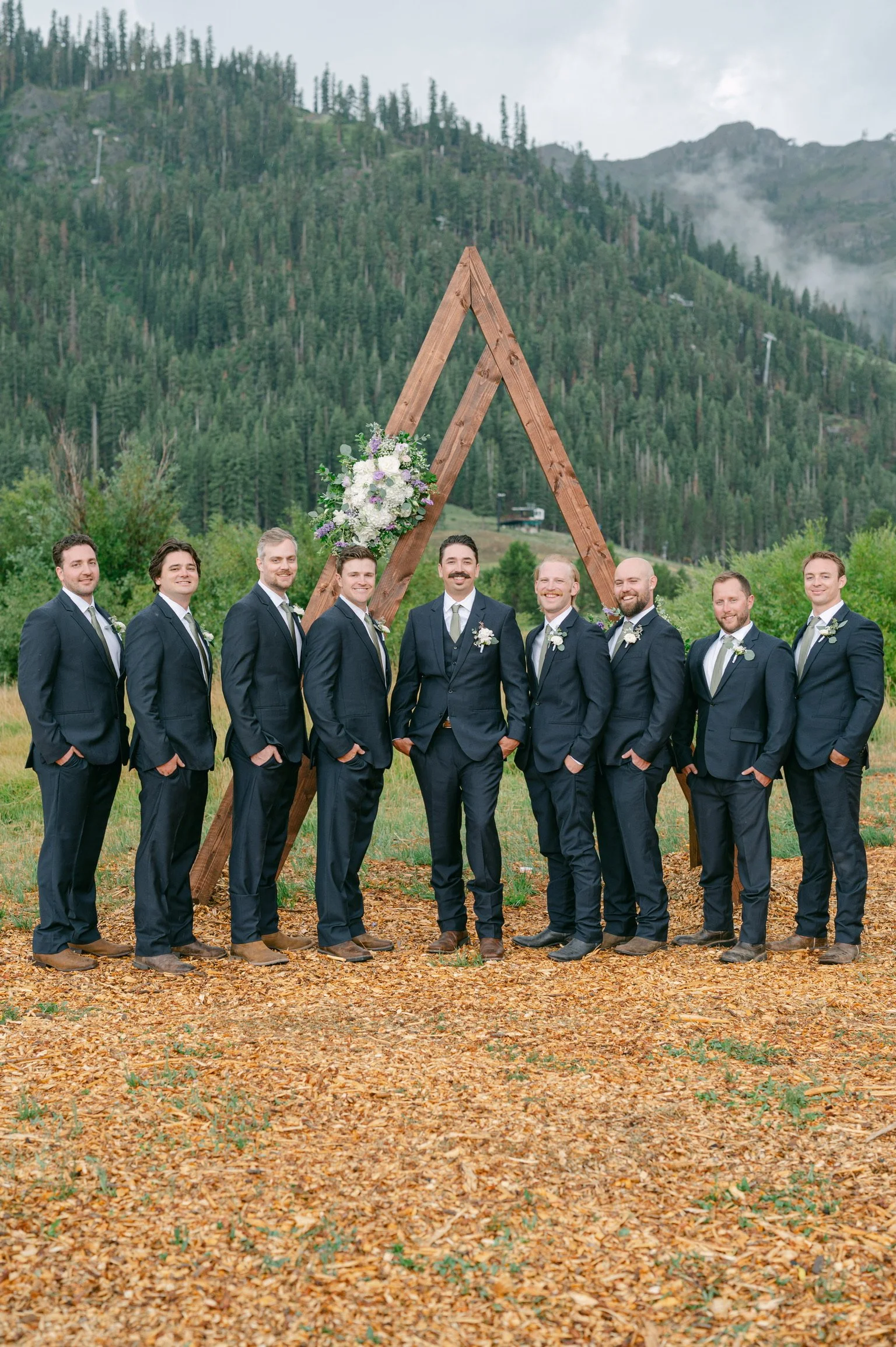 A group of nine men in suits standing outdoors in front of a wooden wedding arch decorated with flowers, with forested mountains in the background.