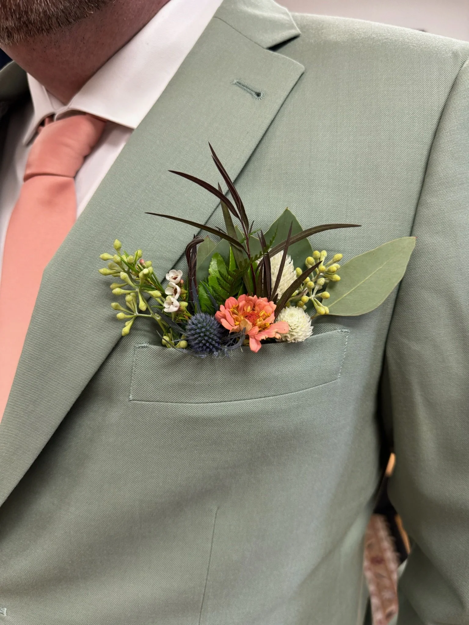 Close-up of a person's green suit with a light pink boutonniere on the lapel, wearing a white shirt and a pink tie.