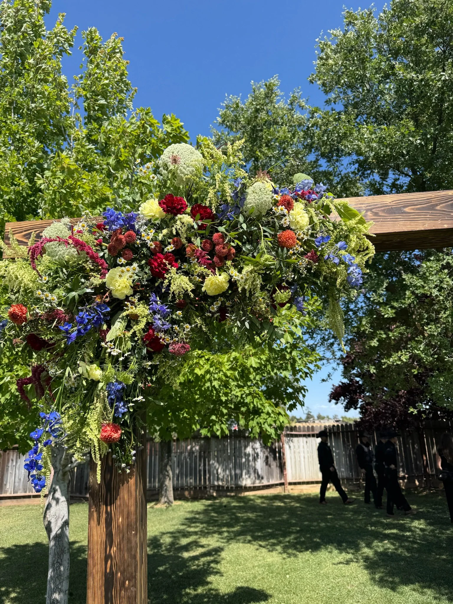 Colorful flower arrangement hanging from wooden posts outdoors under a clear blue sky, with trees and a wooden fence in the background, and some people walking in the yard.