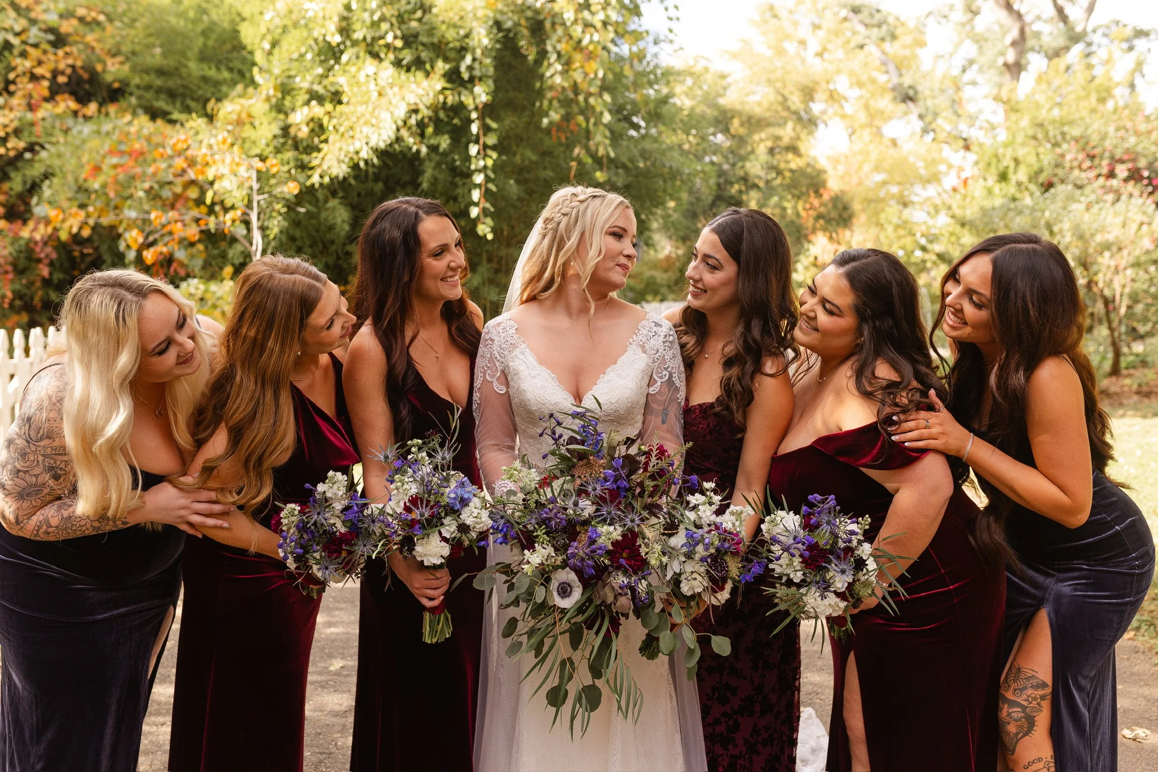 A group of women, including a bride in a white lace wedding dress and veil, surrounded by bridesmaids in burgundy velvet dresses, posed outdoors with trees showing fall foliage.