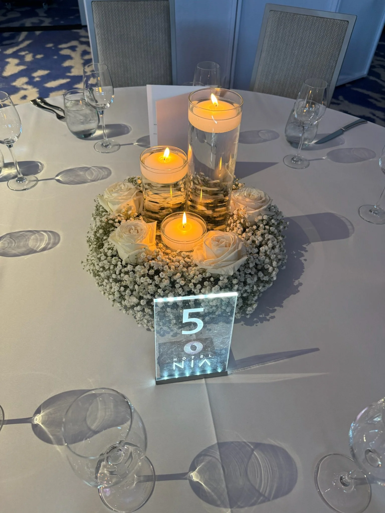 A round dining table with a floral centerpiece featuring white roses and baby's breath, surrounded by empty wine glasses, with three tall floating candles in glass holders and a hotel table number card displaying the number five.