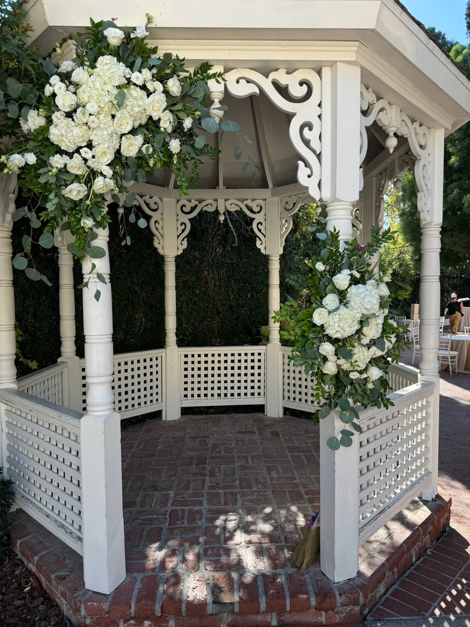White decorative gazebo with intricate scrollwork, adorned with white floral arrangements including roses, hydrangeas, and greenery, situated outdoors on a brick patio.