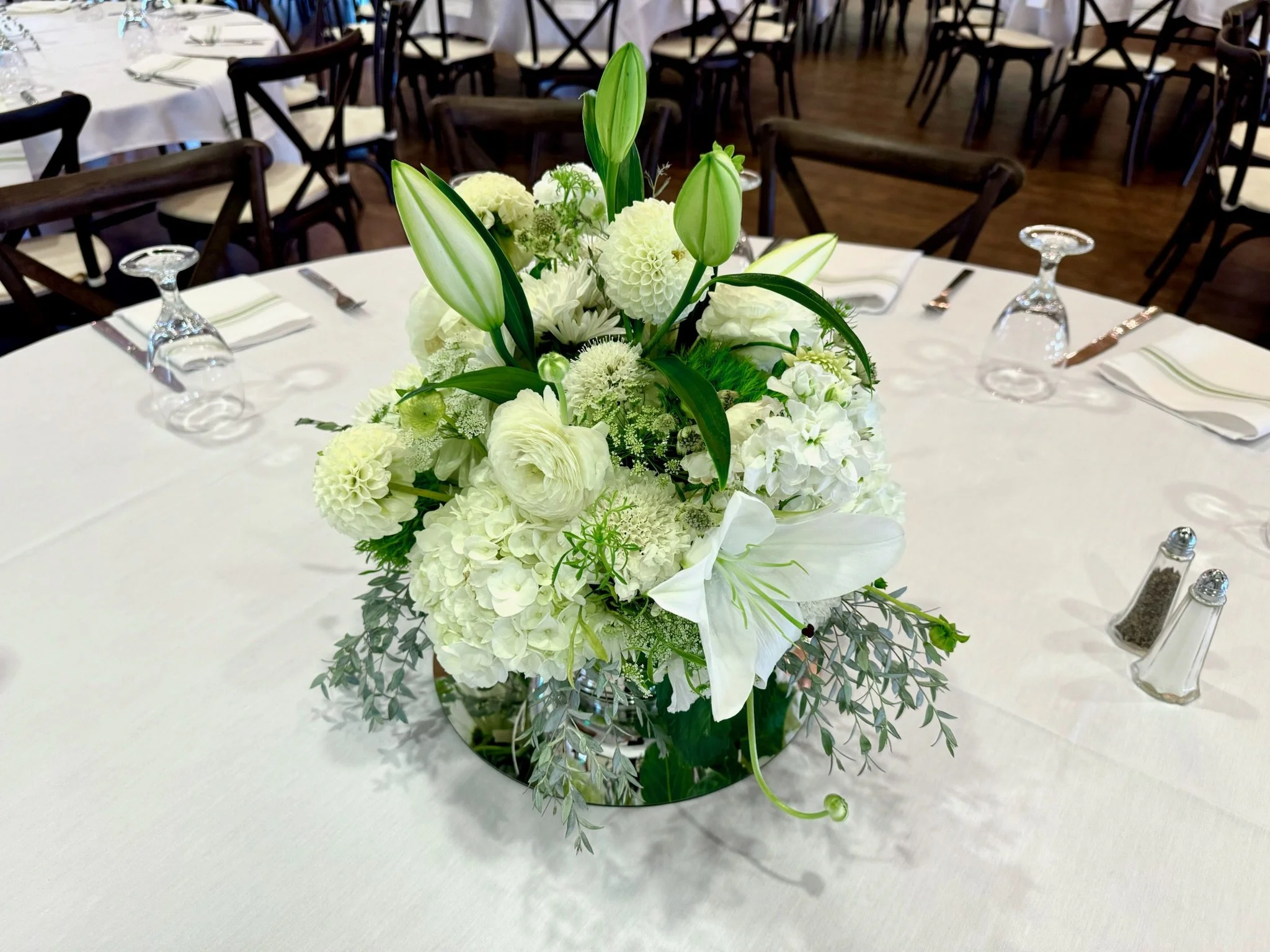 A white floral centerpiece with lilies, dahlias, and greenery on a table set for an event.