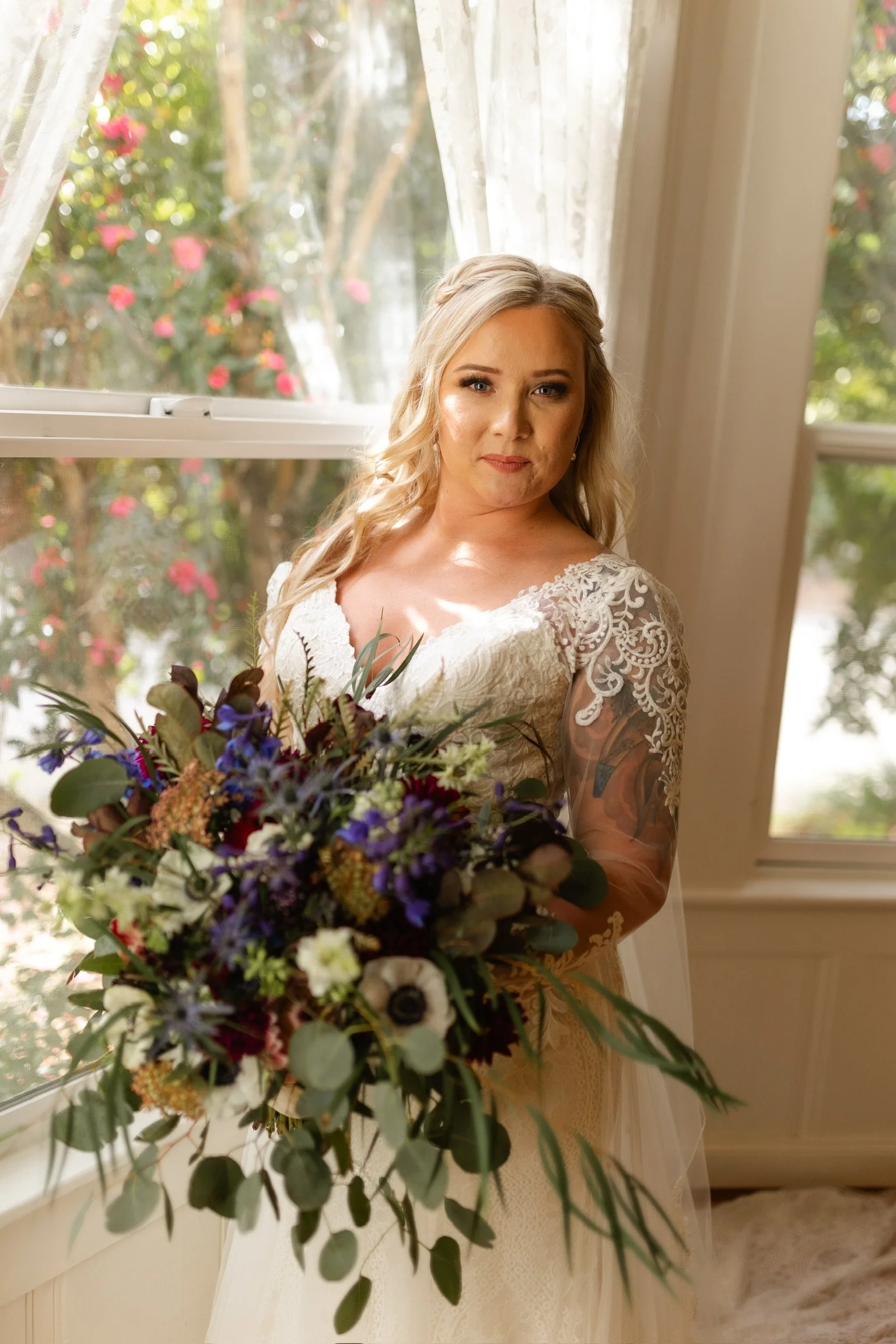 A woman in a lace wedding dress holding a large bouquet of colorful flowers, standing near a window with lace curtains, sunlight shining through, with trees and pink flowers visible outside.