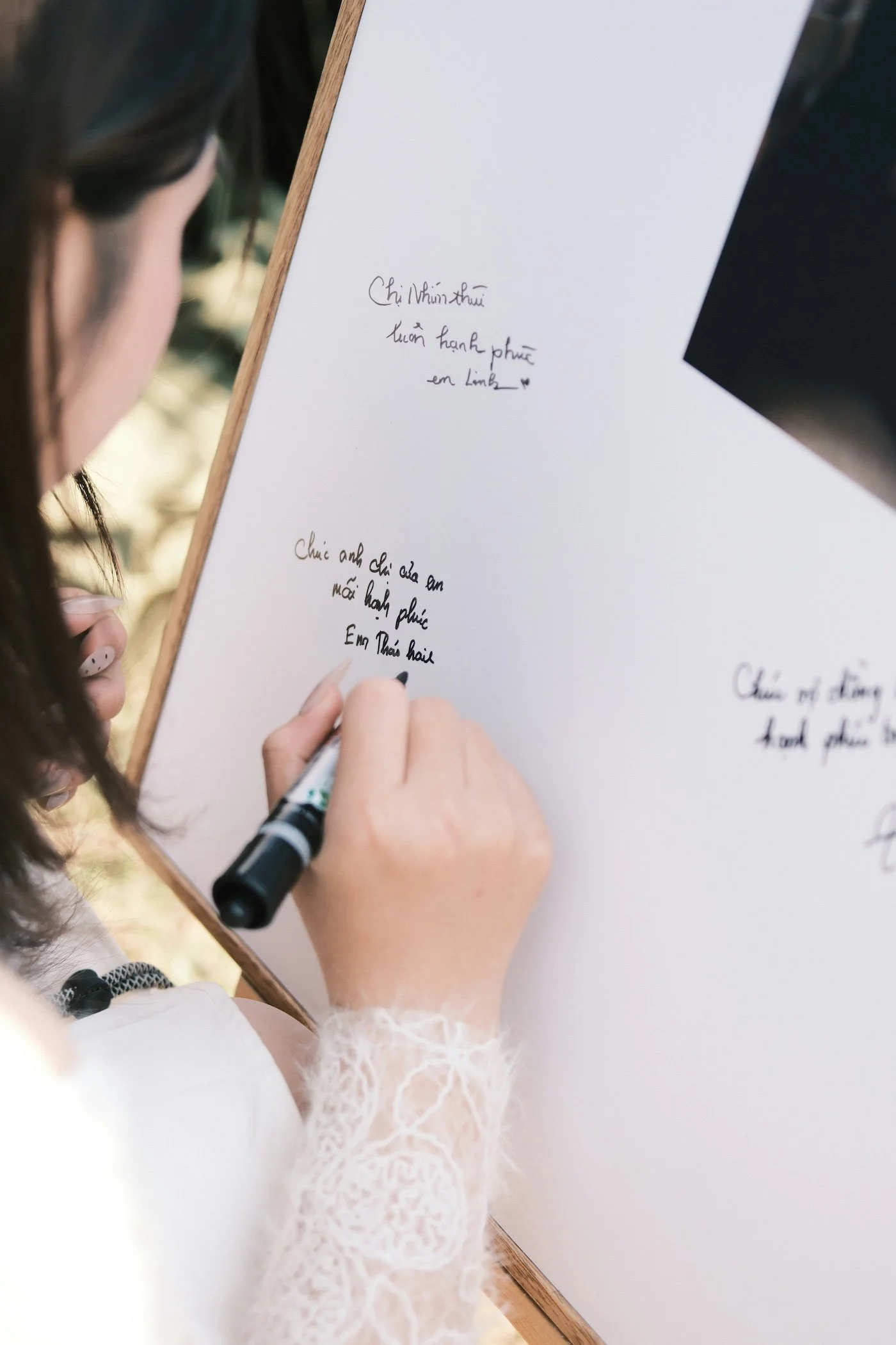 A woman in a white lace sleeve writes on a white paper with Vietnamese handwritten text, holding a black marker. Part of her face and hand are visible, and the paper is on a wooden surface.