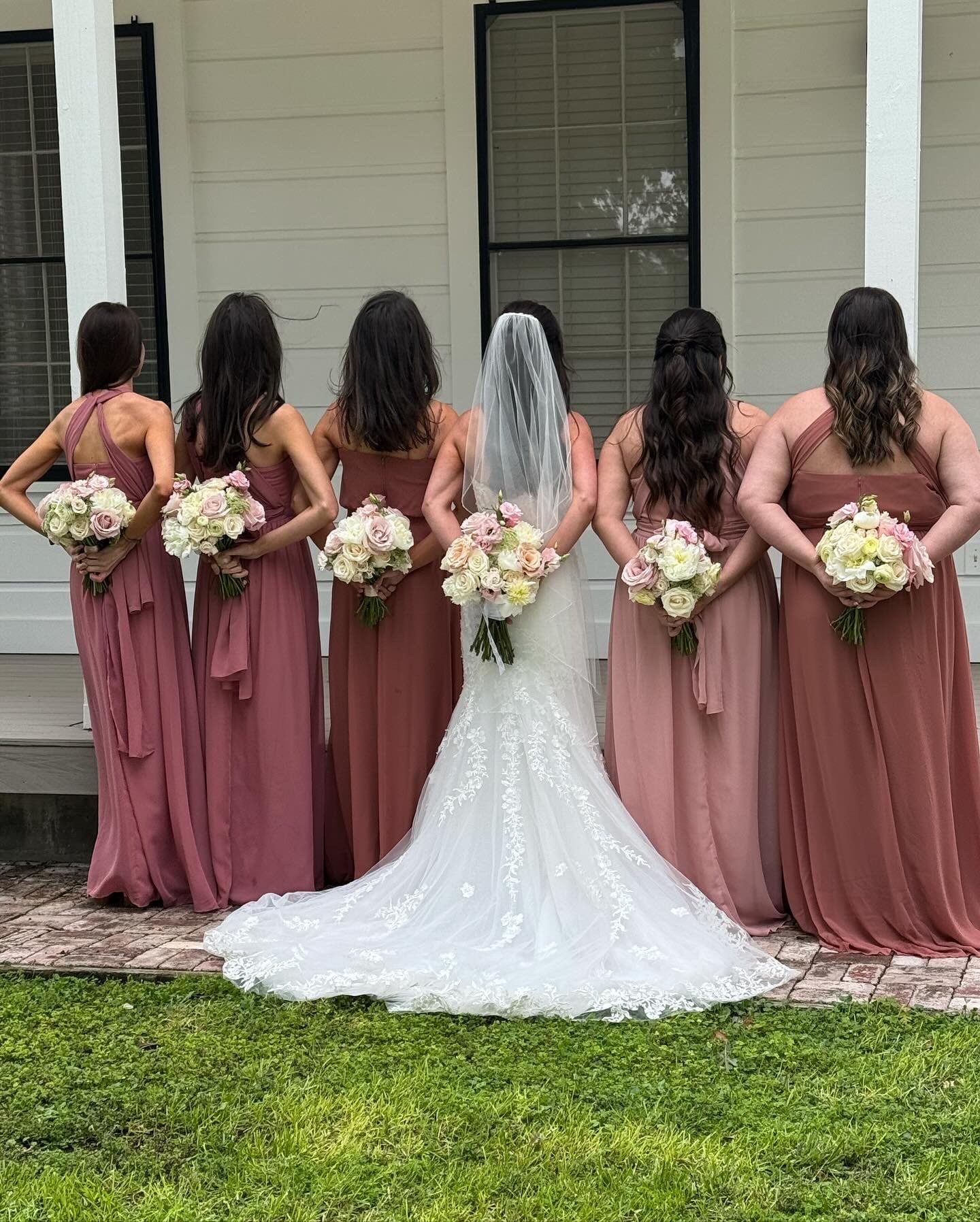 Six women in matching mauve bridesmaid dresses standing with their backs to the camera, each holding a bouquet of pink and white roses. The bride, in a white wedding gown with a long lace train and veil, stands in the center with her back to the came