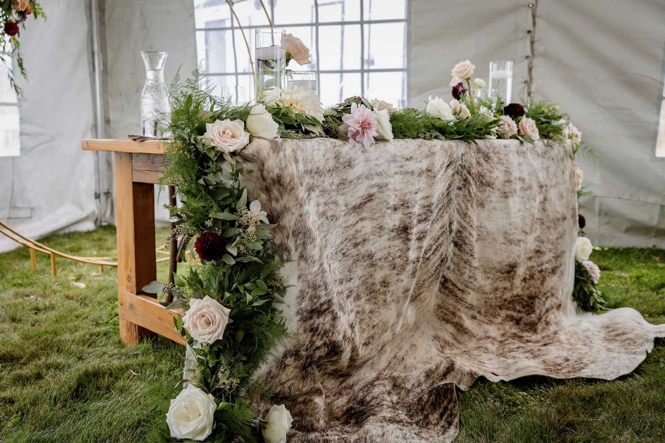 A floral wedding table arrangement with white roses, pink dahlias, greenery, and candles on a draped fabric tablecloth inside a tent with a windowed background.