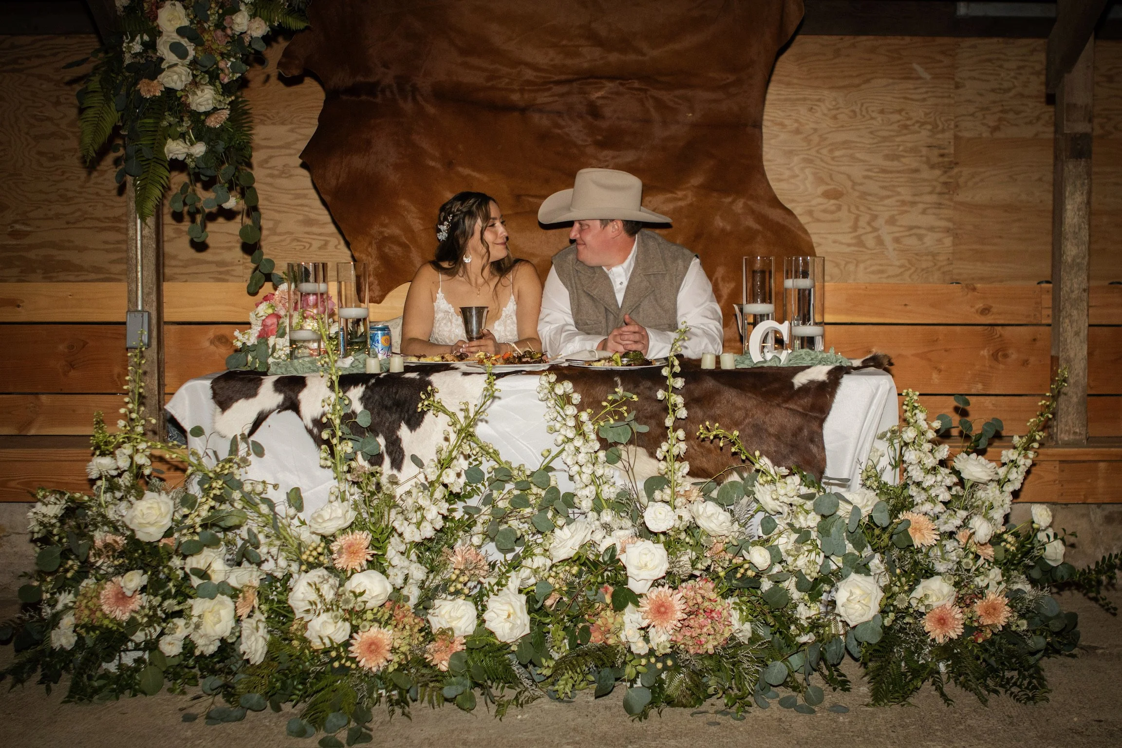 A bride and groom sitting at a decorated wedding reception table, facing each other and smiling, surrounded by floral arrangements and greenery.