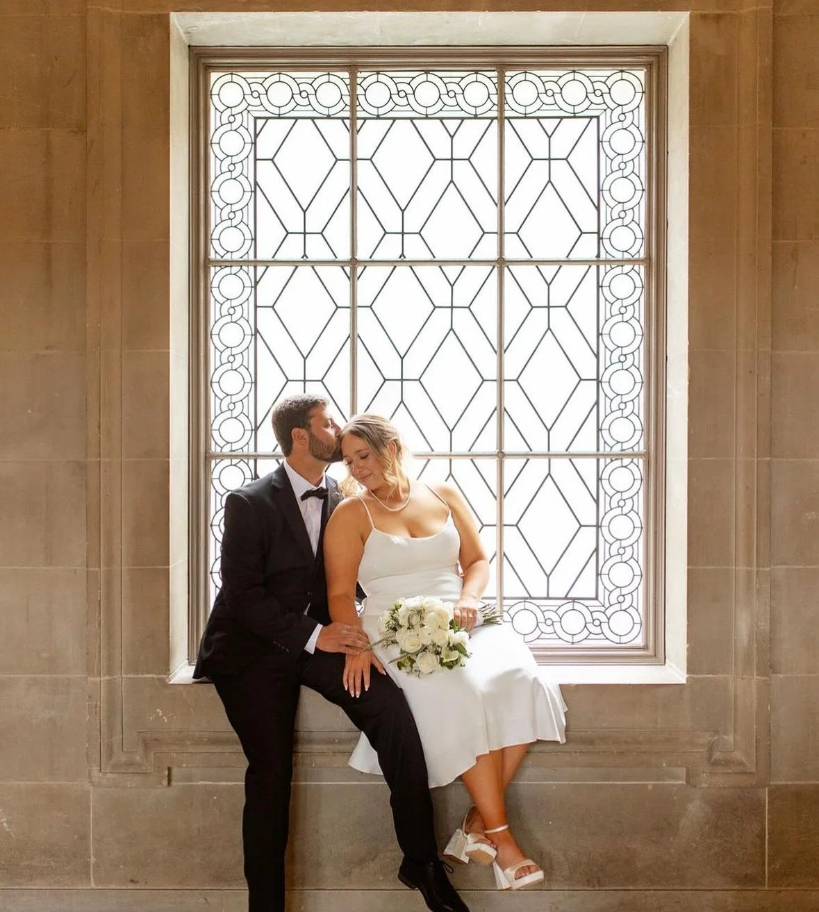 A bride and groom sitting together in front of a large decorative window, with the groom kissing the bride on the side of her head. The bride is holding a bouquet of white flowers, and they are both dressed in formal wedding attire.