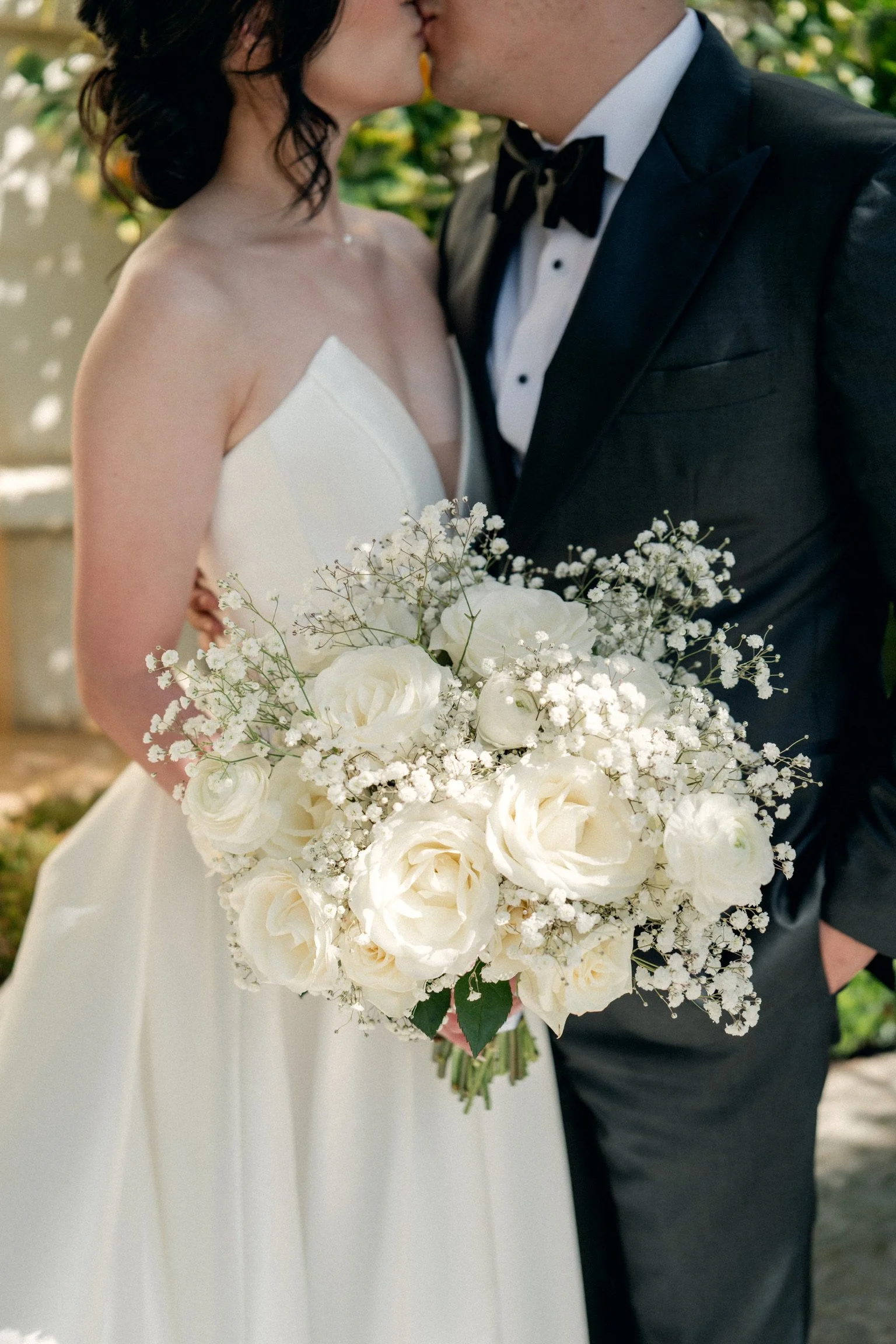 A bride and groom kissing, with the bride holding a large bouquet of white roses and baby's breath, outdoors with greenery in the background.