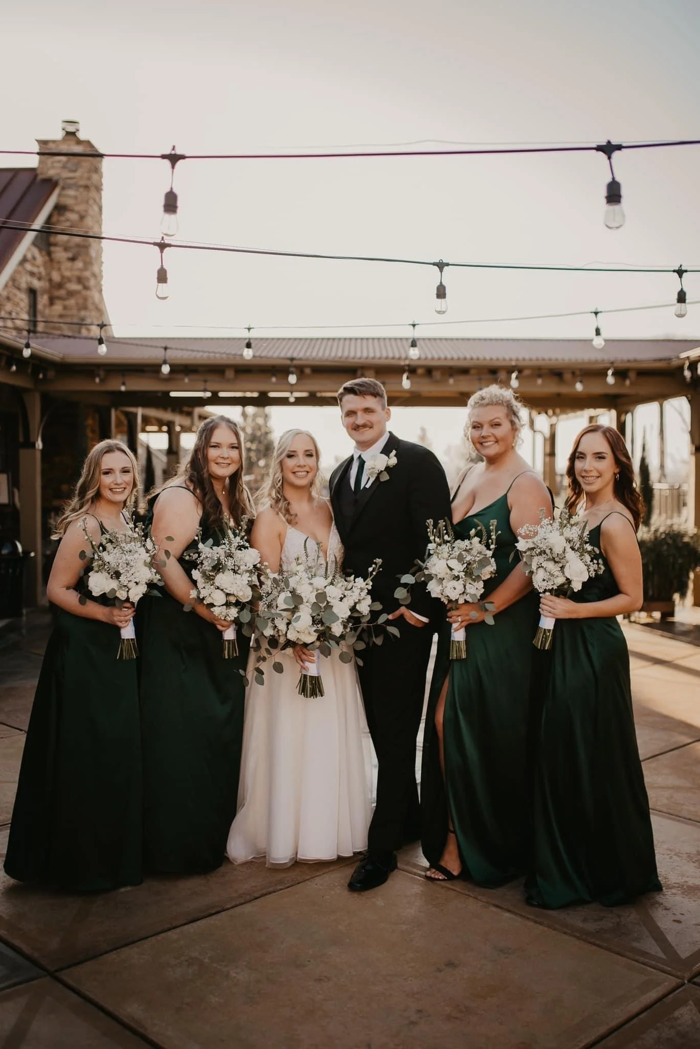 Group of wedding bride and bridesmaids with the groom at an outdoor venue during sunset, holding bouquets of white flowers and dressed in formal attire.