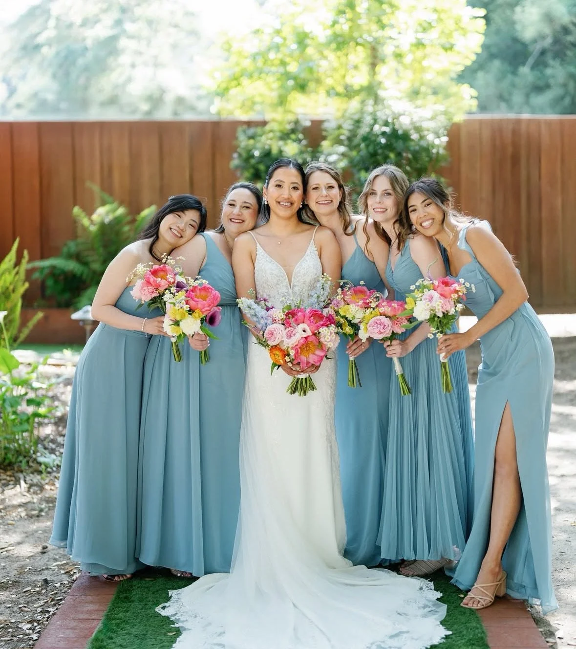 A bride in a white wedding dress standing with five bridesmaids in light blue dresses, holding bouquets of pink and white flowers outdoors with a wooden fence and green trees in the background.