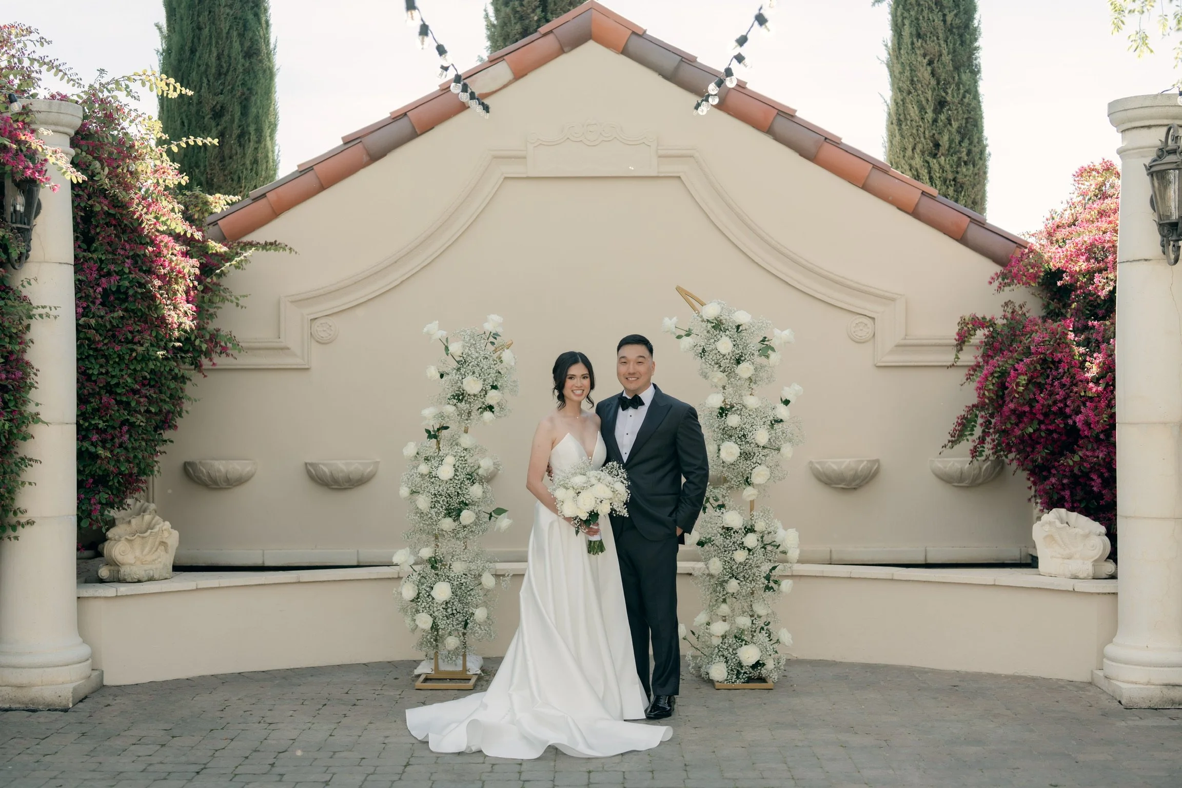 A newlywed couple stands outside in front of a white wall decorated with flowers. The bride is in a white wedding gown holding a bouquet, and the groom is in a black tuxedo with a bow tie. They are smiling at the camera under string lights. The setti