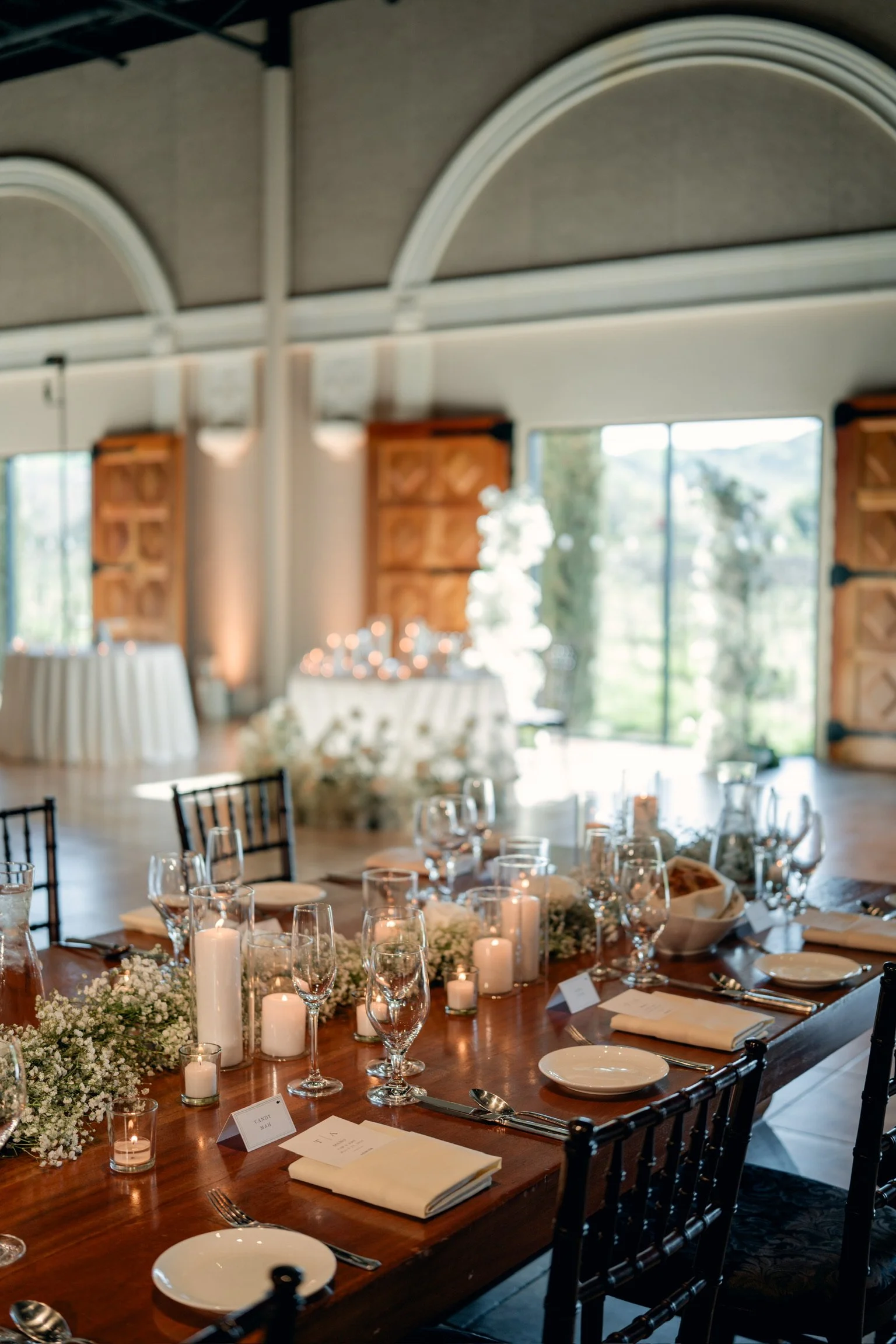 Indoor event space decorated with tables set for a formal gathering, with wine glasses, candles, and floral arrangements, large windows letting in natural light, and exposed wooden beams on the ceiling.