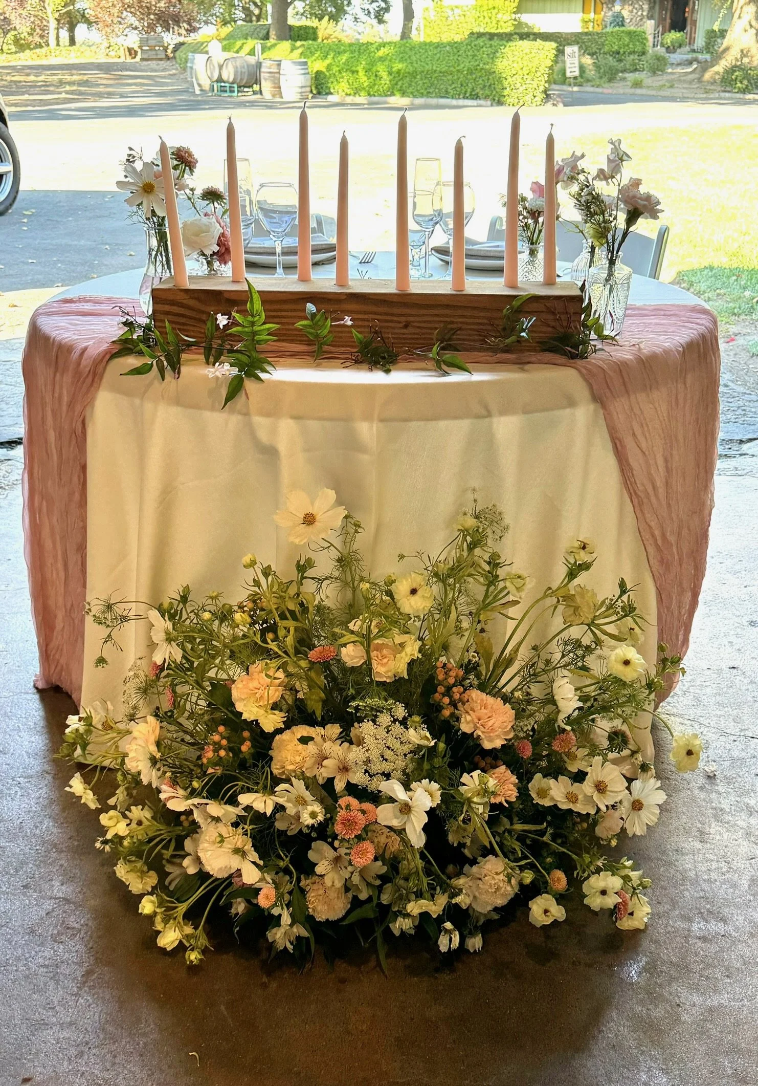 A table decorated with pink cloth and surrounded by flowers, a wooden candle holder with light pink candles, and small flower arrangements in vases in a bright setting.