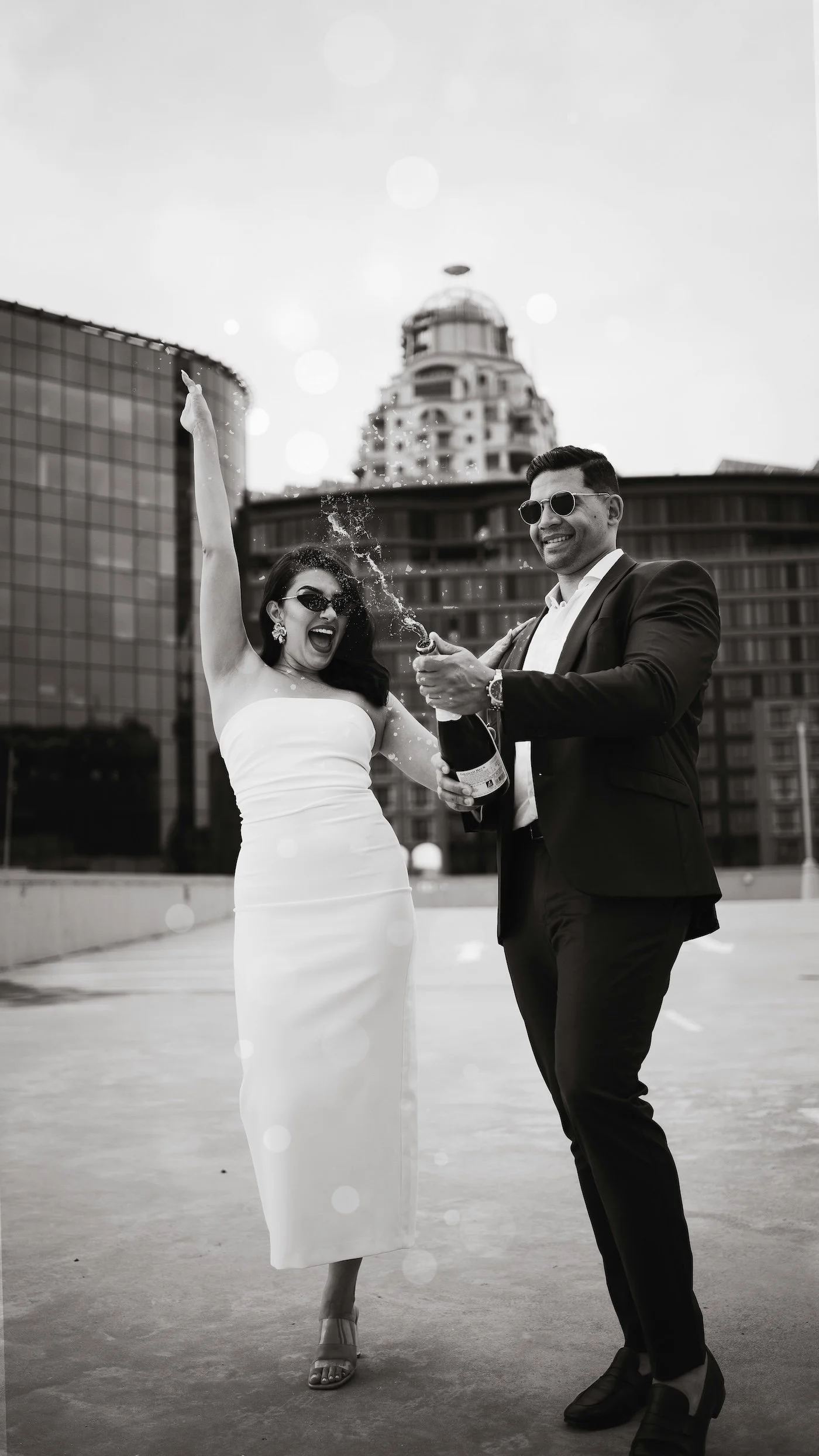 A joyful woman in a strapless dress and sunglasses celebrating with a man in a suit and sunglasses, opening a bottle of champagne with a city skyline in the background.