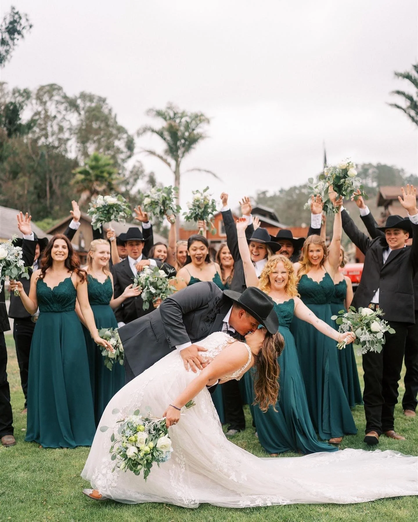 A wedding celebration outdoors with a bride and groom kissing, surrounded by bridesmaids and groomsmen, some holding bouquets and raising their hands in celebration.