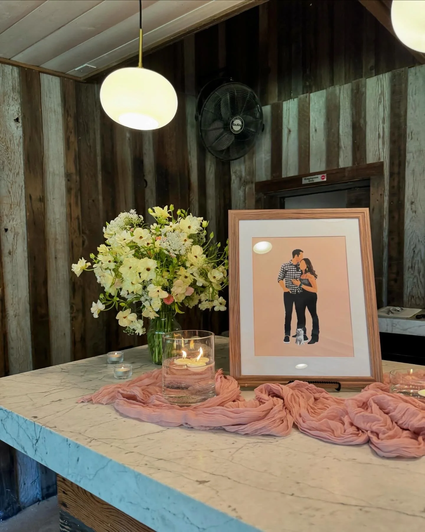 A marble tabletop decorated with a framed illustration of a couple, a bouquet of white flowers, candles, and pink fabric. The background shows a wooden wall, a wall-mounted fan, and a hanging light fixture.