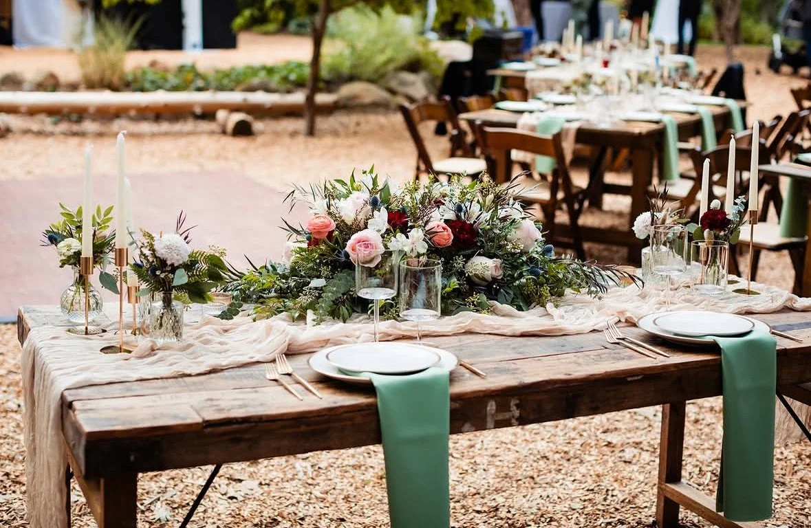 Rustic outdoor wedding table decorated with floral centerpiece, candles, and tableware, set in a natural environment with trees and other tables in background.