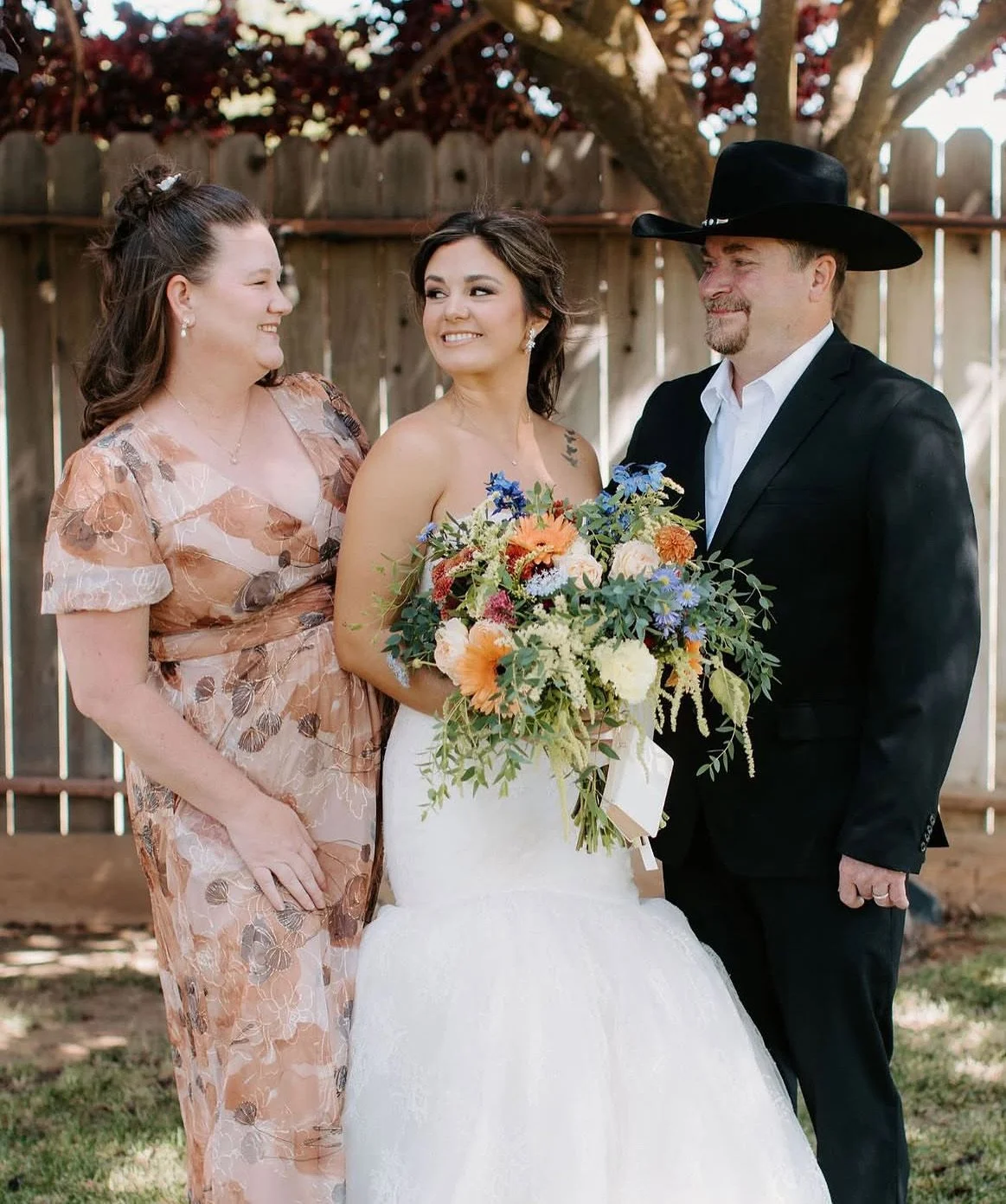 A bride holding a colorful bouquet of flowers, standing between a woman in a floral dress and a man in a black suit and cowboy hat, in an outdoor setting with a wooden fence and trees in the background.