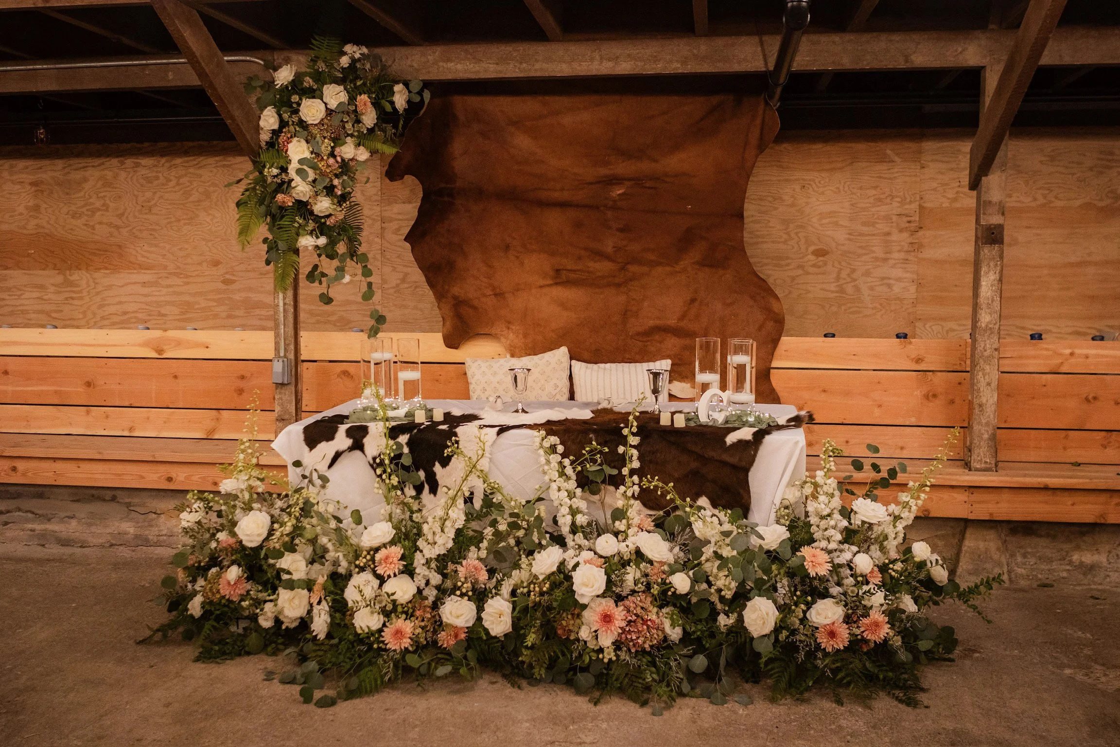 A wedding reception table decorated with white and pink flowers, green foliage, glasses, and candles, against a wooden backdrop with a large, irregularly shaped wooden panel and floral arrangement hanging on the left side.