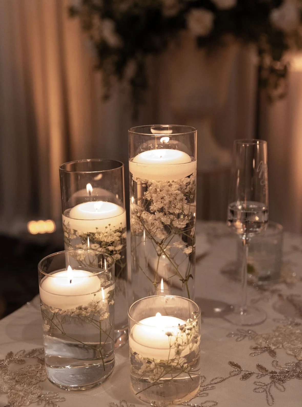Several lit candles in glass holders filled with water and white flowers, on a decorated table with a lace tablecloth, creating an elegant ambiance.
