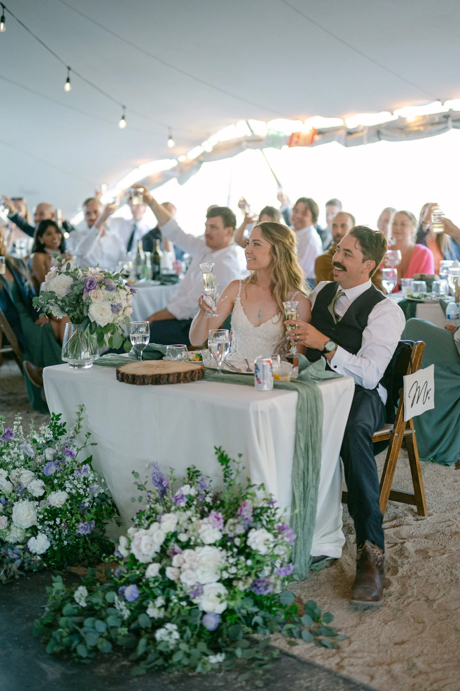 A bride and groom sitting at a table surrounded by floral decorations, raising glasses in a toast at their wedding reception.