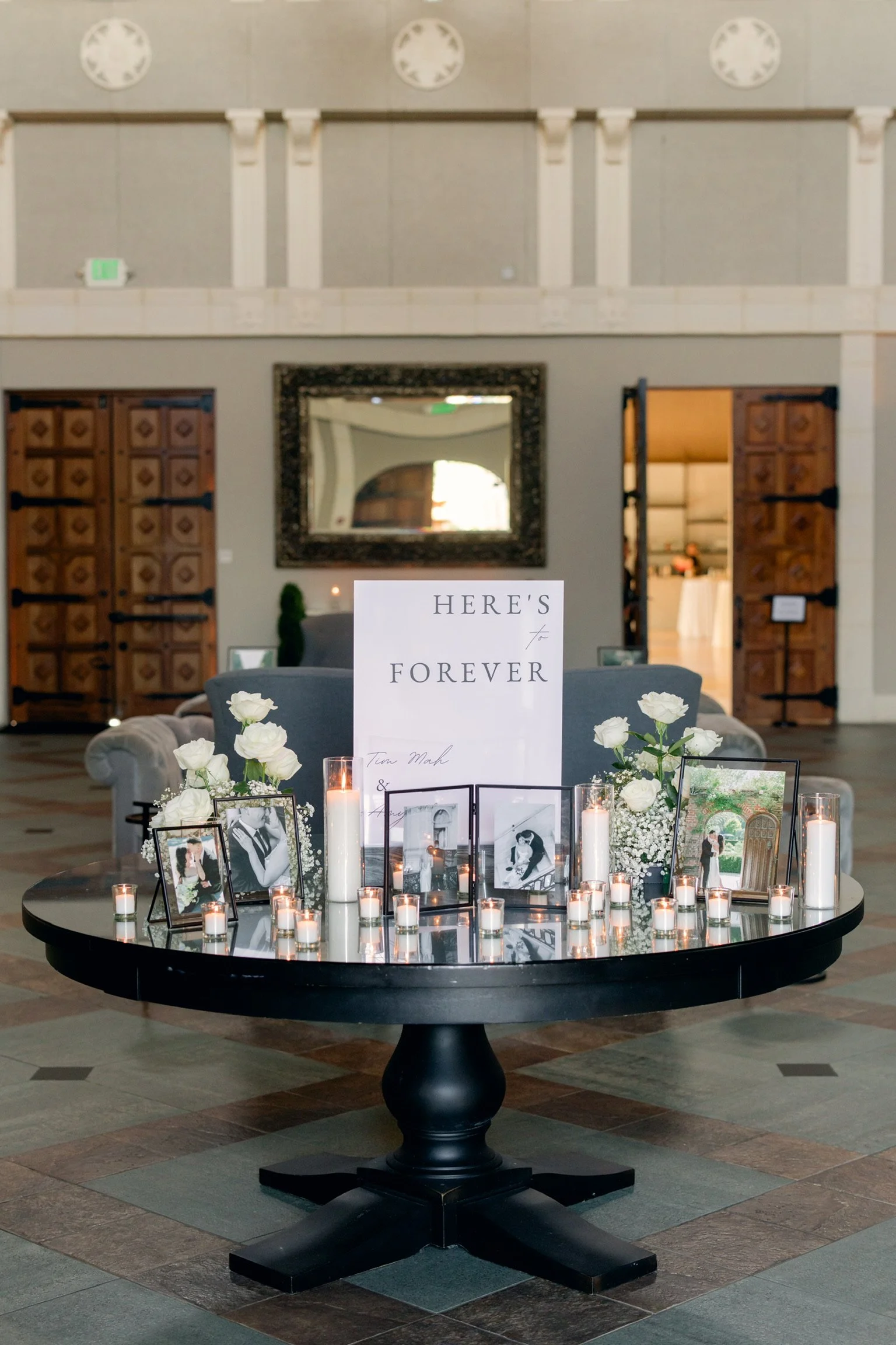 A round black table decorated with white flowers, candles, framed photos, and a sign that reads "Here's Forever" at a wedding or memorial event inside a spacious hall with high ceilings, wall clocks, and wooden doors.
