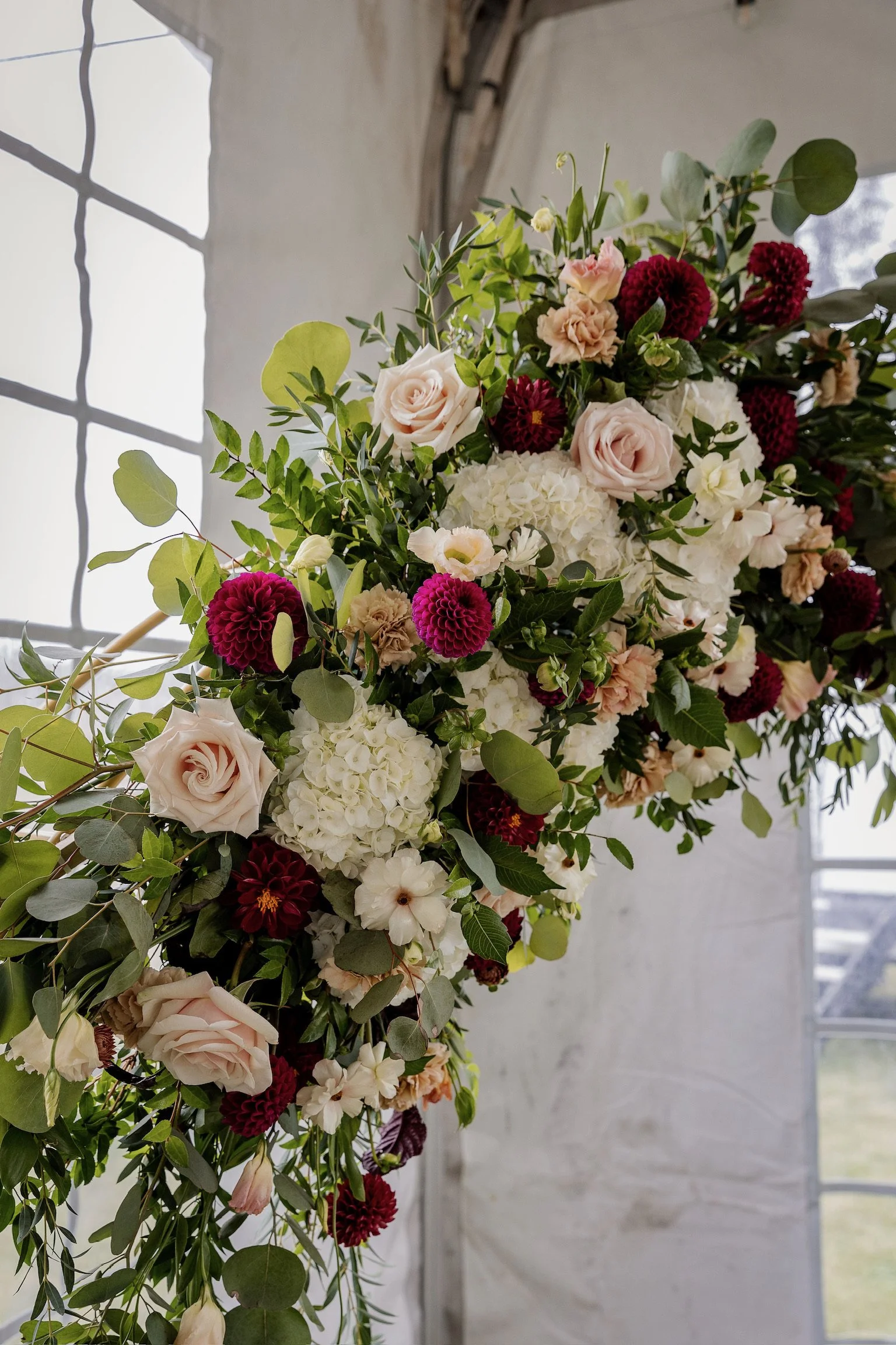 A large, colorful floral arrangement with roses, hydrangeas, and other flowers, set against a white brick wall with windows in the background.