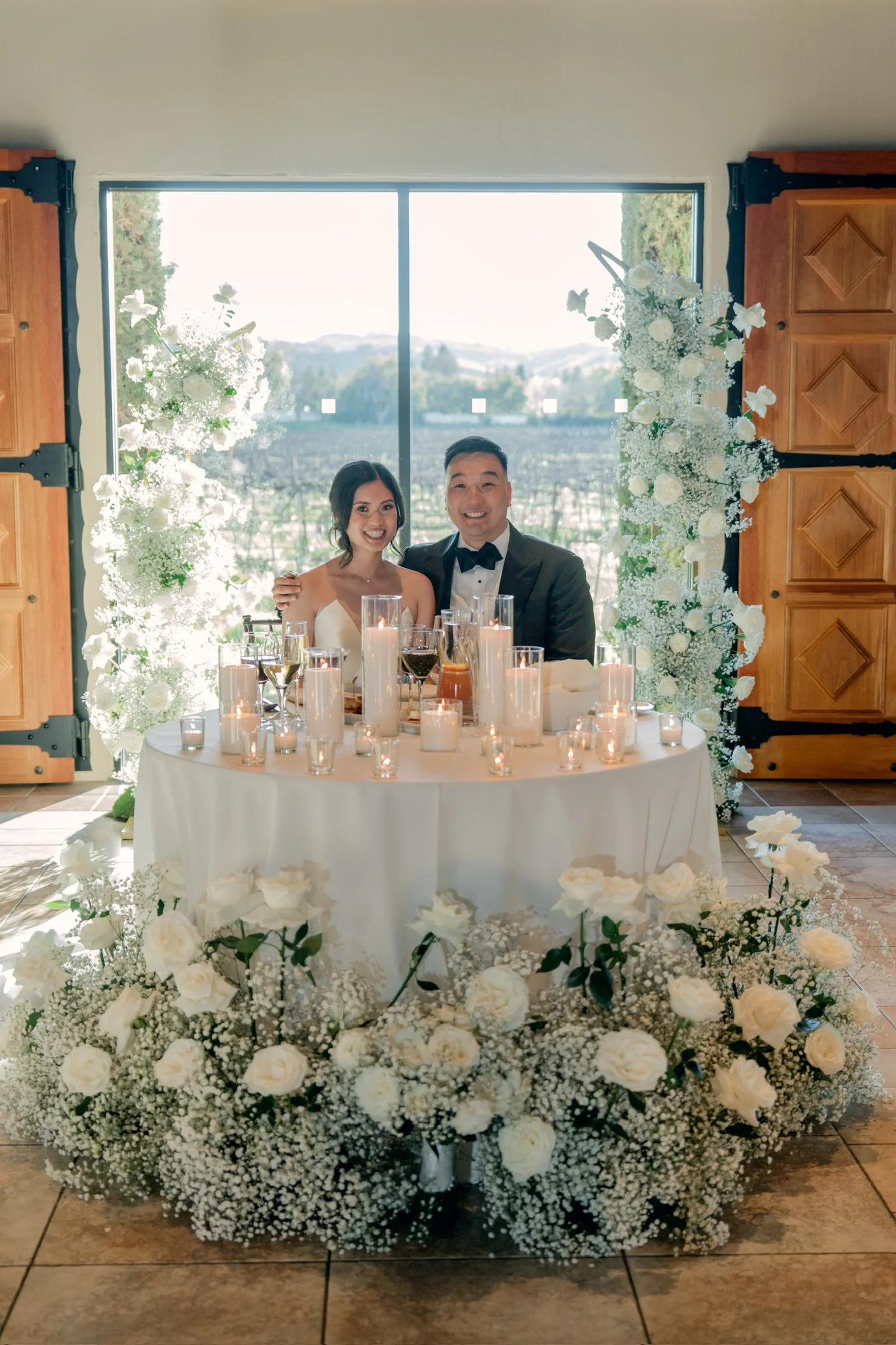 A smiling couple at a wedding reception, sitting at a decorated table with candles, surrounded by white flowers, with large windows and a scenic outdoor view in the background.
