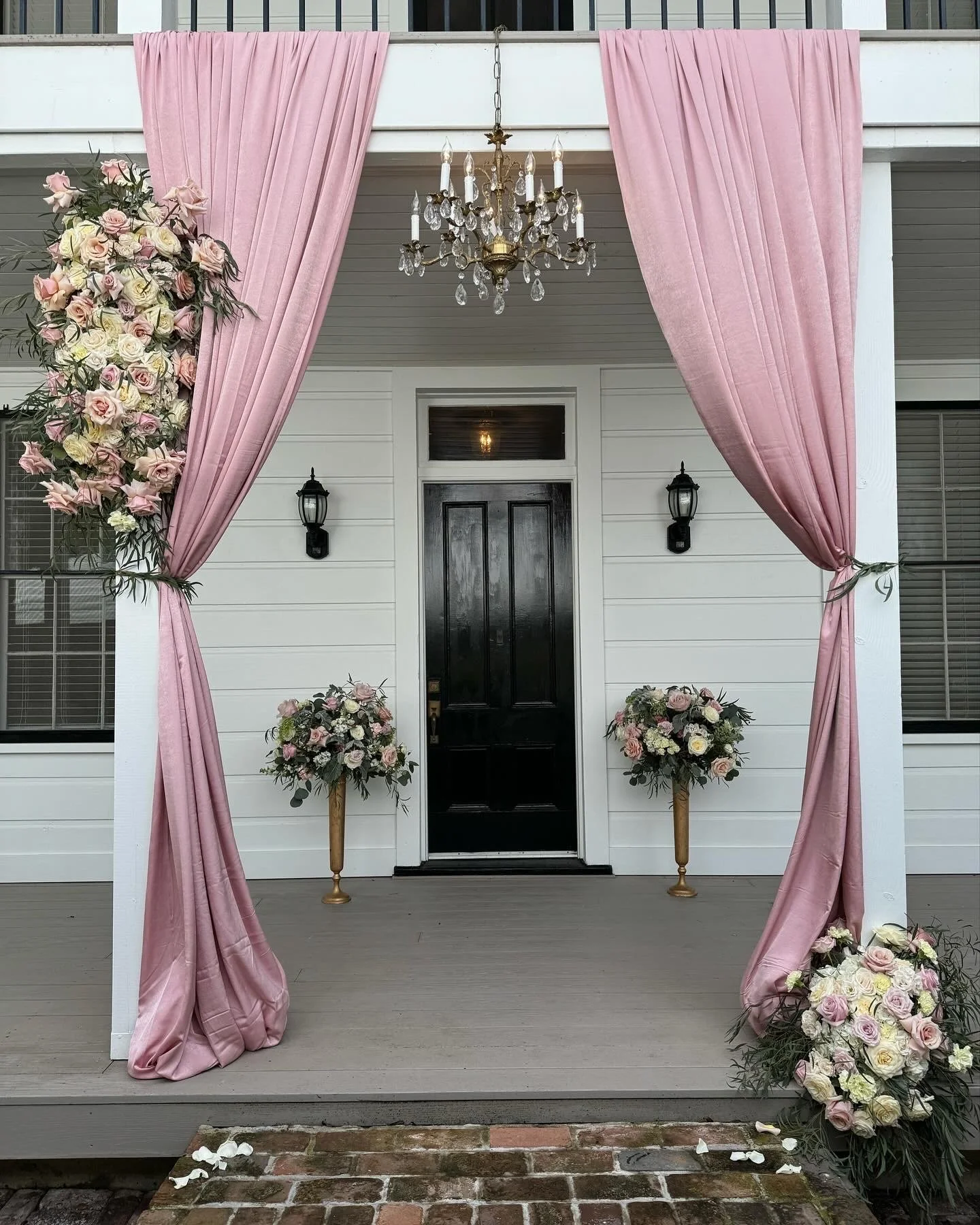 A decorated front porch with pink curtains tied back, floral arrangements on either side, a black front door, and a chandelier hanging from above.