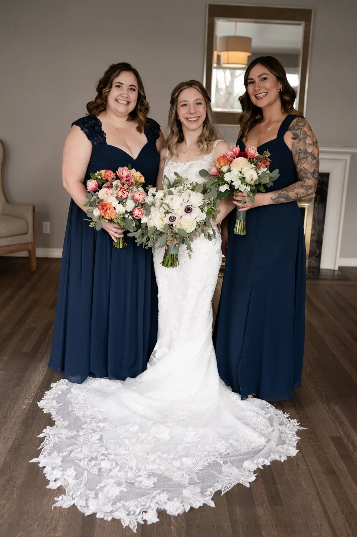 Three women, one bride in a white wedding dress, flanked by two bridesmaids in navy blue dresses, all holding colorful bouquets, standing indoors in front of a mirror and fireplace.