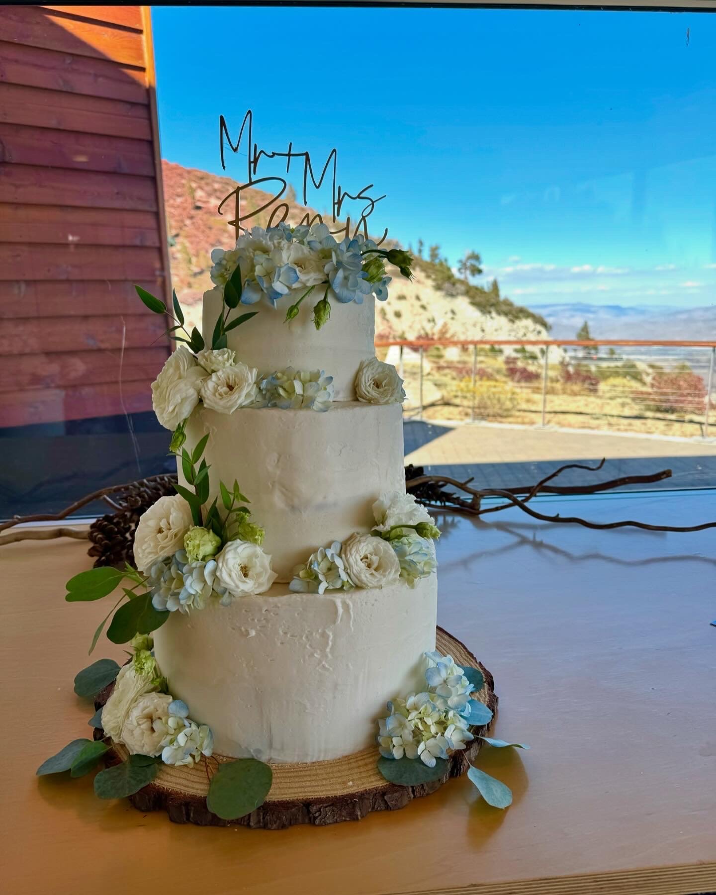 A three-tier white wedding cake decorated with white and light blue flowers, green leaves, and a gold 'Mom & Mrs. Pam' cake topper, placed on a wooden slab with a scenic mountain view in the background.