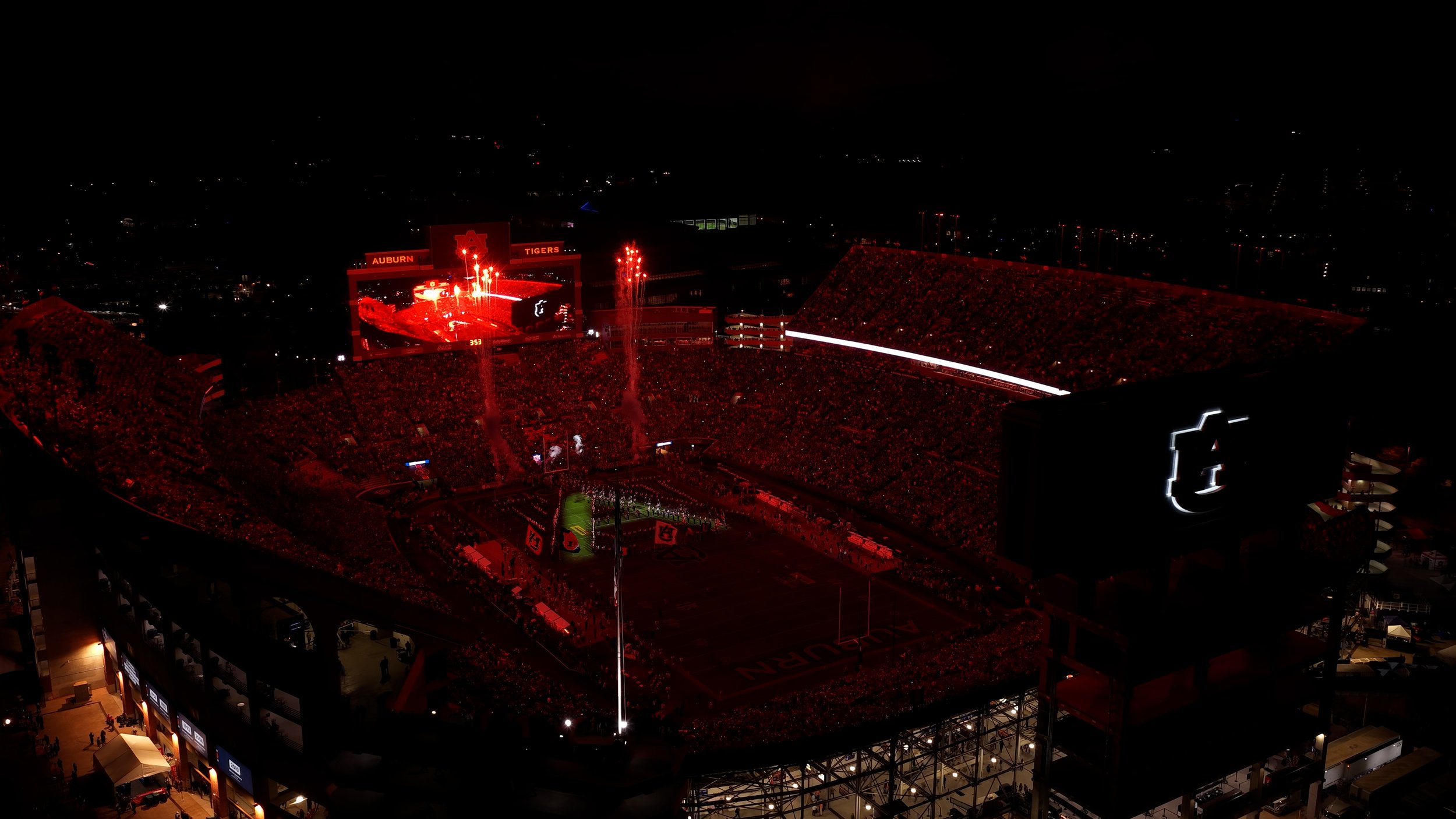 Nighttime view of a football stadium lit in red with fireworks, crowd, and large screens displaying the Auburn Tigers logo and games, indicating a game or event.