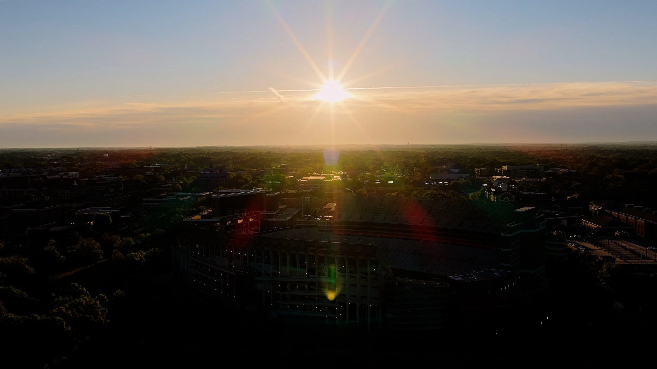 Sunset over a city with buildings, trees, and a stadium in the foreground, bright sunlight creating lens flares.