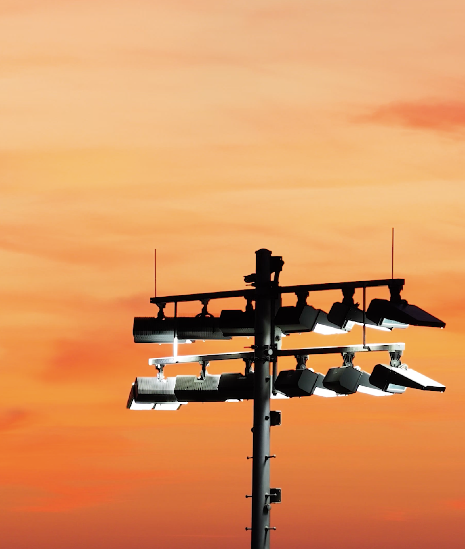 A tall stadium floodlight pole with multiple lights against a colorful orange sunset sky.